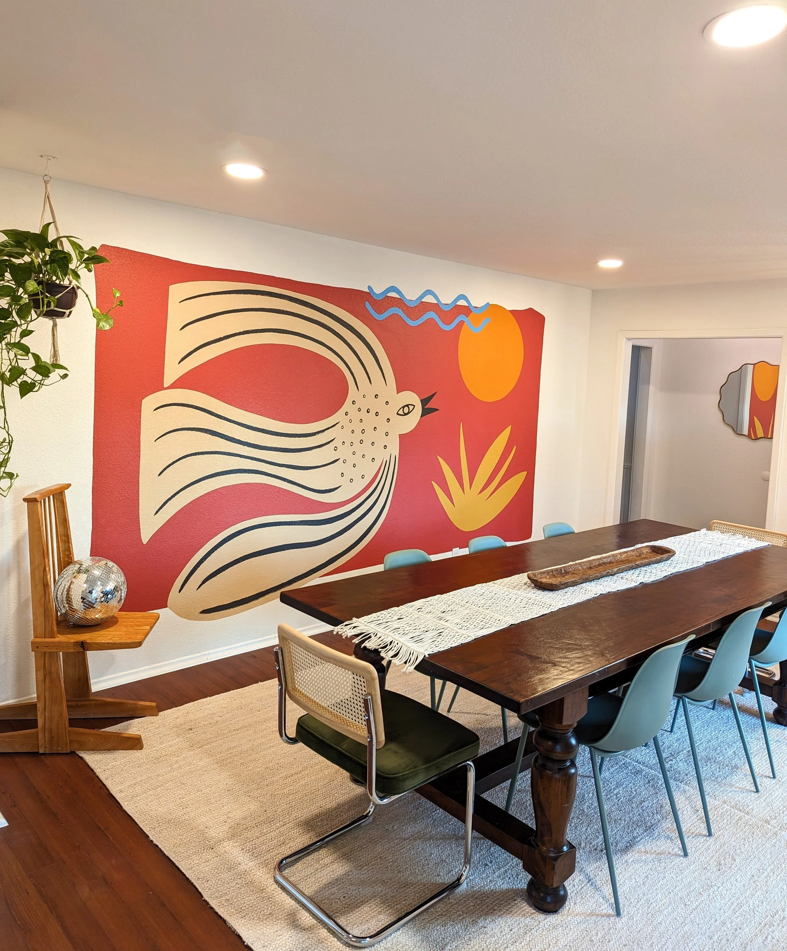 A dining room with a wooden table, colorful chairs, a beige rug, a large abstract wall art, a hanging plant, and ceiling lights.