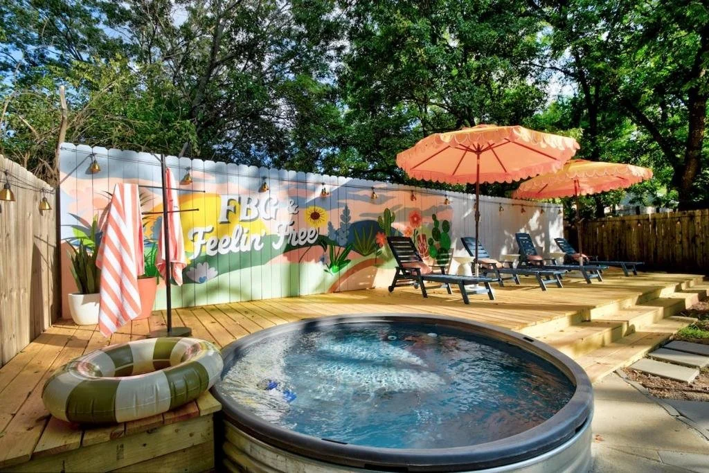 A backyard pool area with a hot tub, patio chairs with umbrellas, and a colorful mural on the fence that says "FBC Feelin' Free" with floral and nature designs, surrounded by trees.