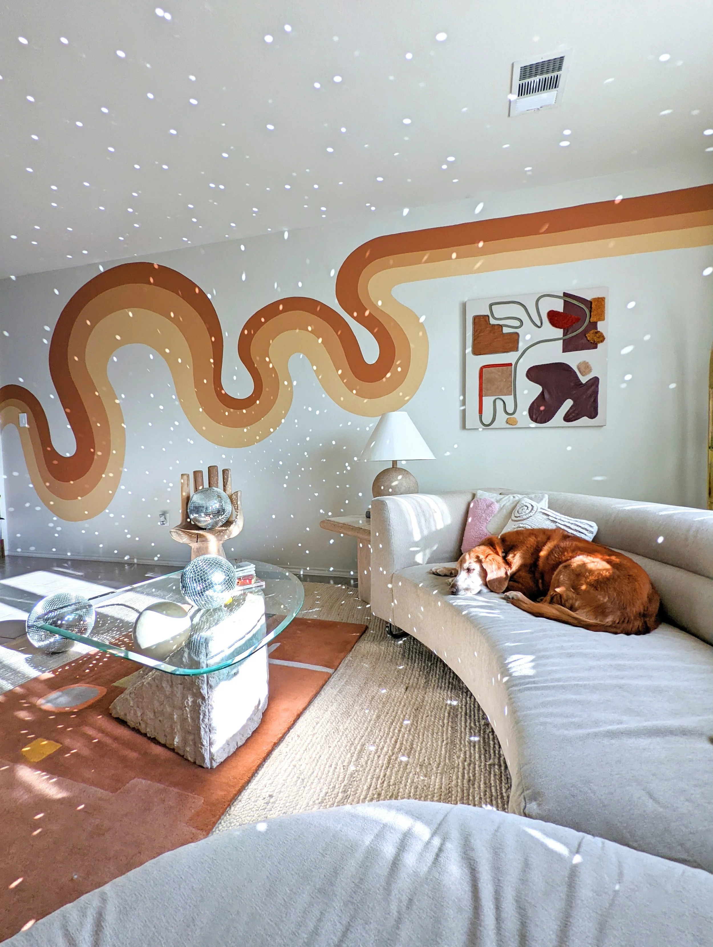 Funky bright and colorful living room in an Austin-area home featuring original artwork, eclectic vintage decor, layered rugs, and a retro wavy wall mural, featured in Apartment Therapy.