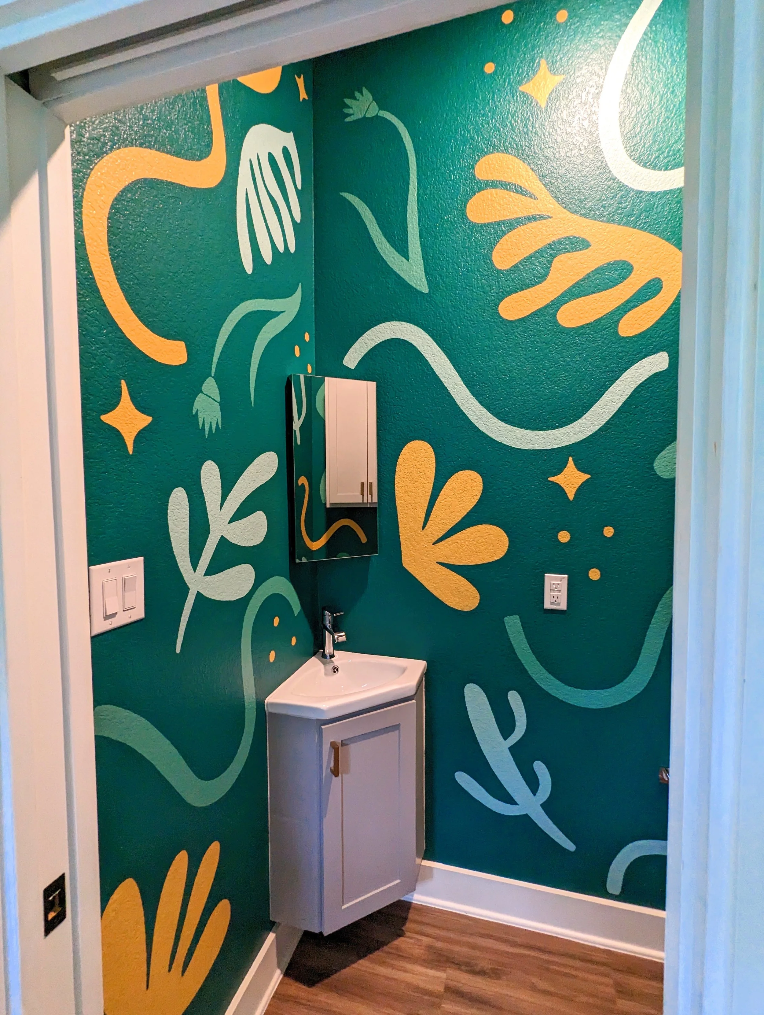 A bathroom corner with teal walls decorated with abstract floral and star patterns in yellow and white, featuring a small white sink with wooden cabinet and a mirror above it, and wood flooring.