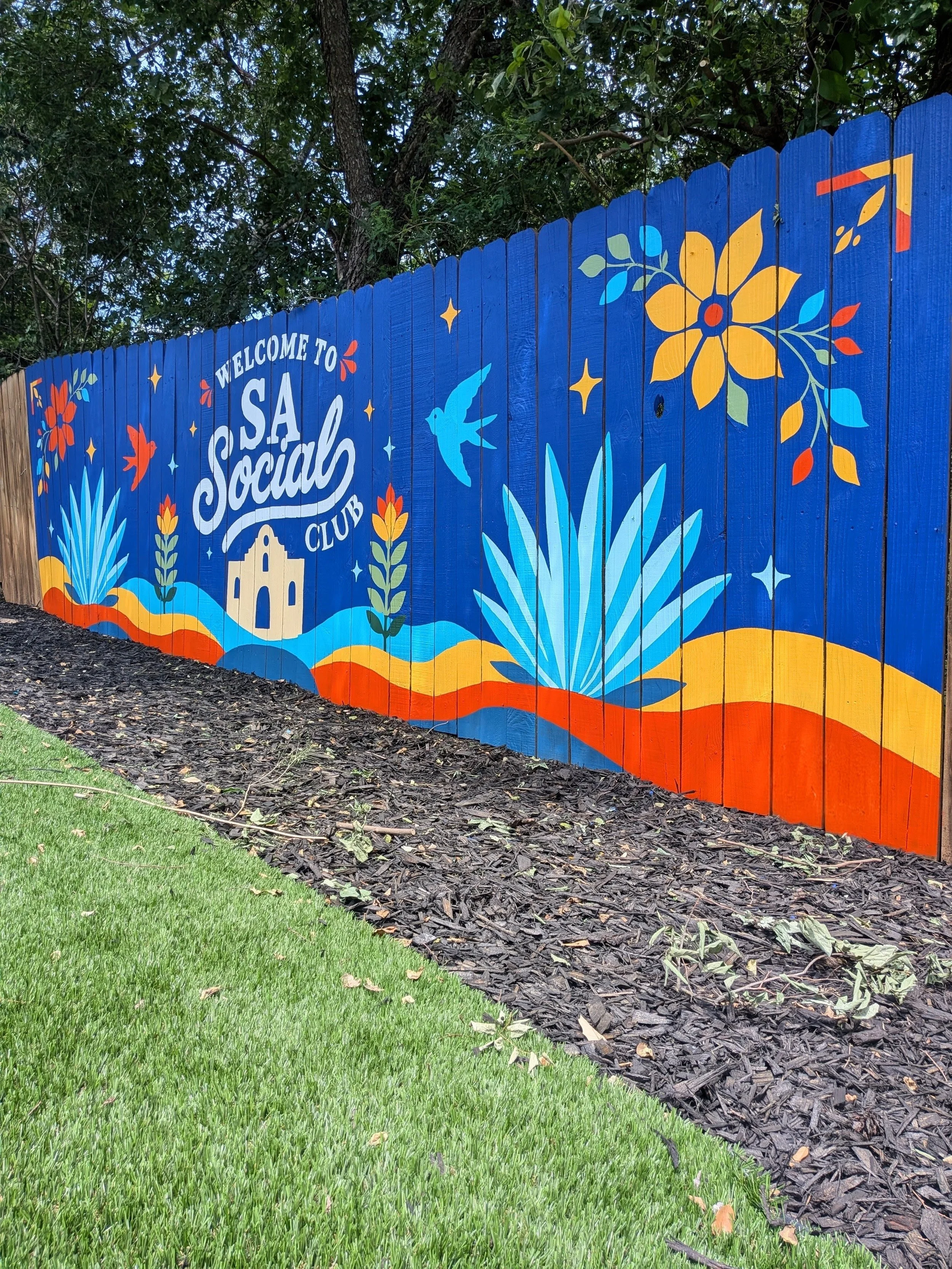 Exterior fence mural for a San Antonio short-term rental featuring the "SA Social Club" logo, Texas flowers, birds, and native plants.