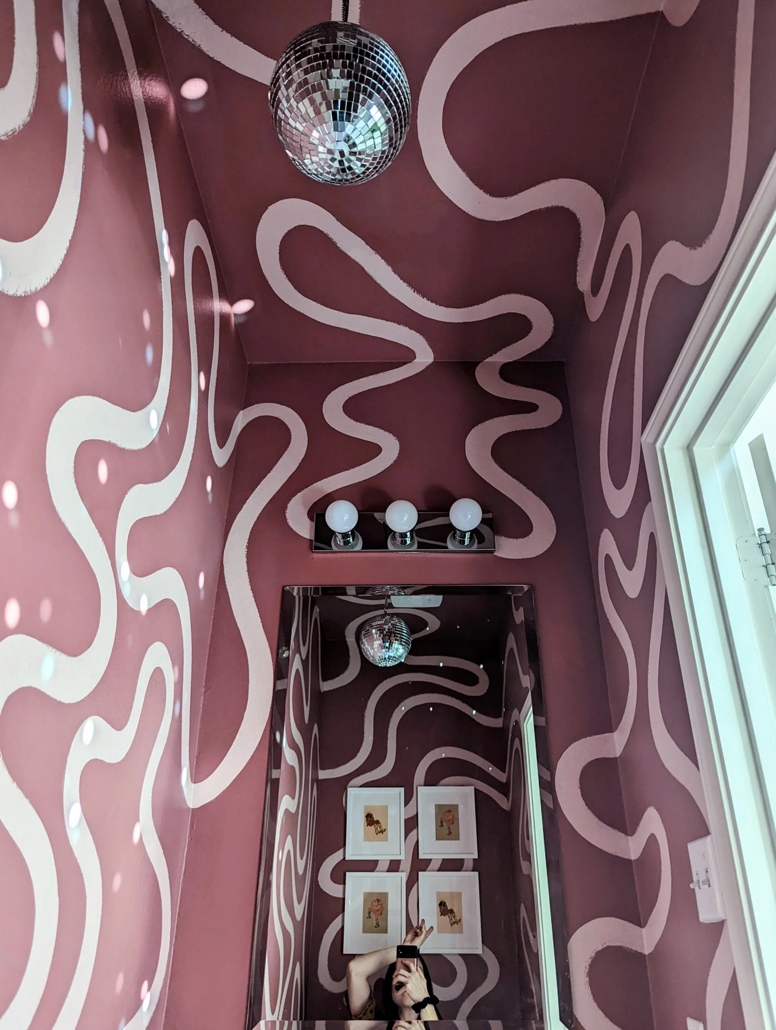 Playful bathroom mural in an Austin-area home featuring mauve purple and white wavy shapes, framed artwork, and a disco ball, featured in Apartment Therapy.