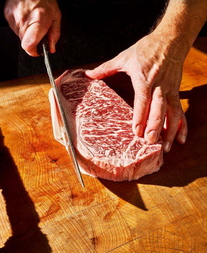 WESTHOLME WAGYU sydney. Person slicing a large, marbled cut of raw beef steak on a wooden cutting board.