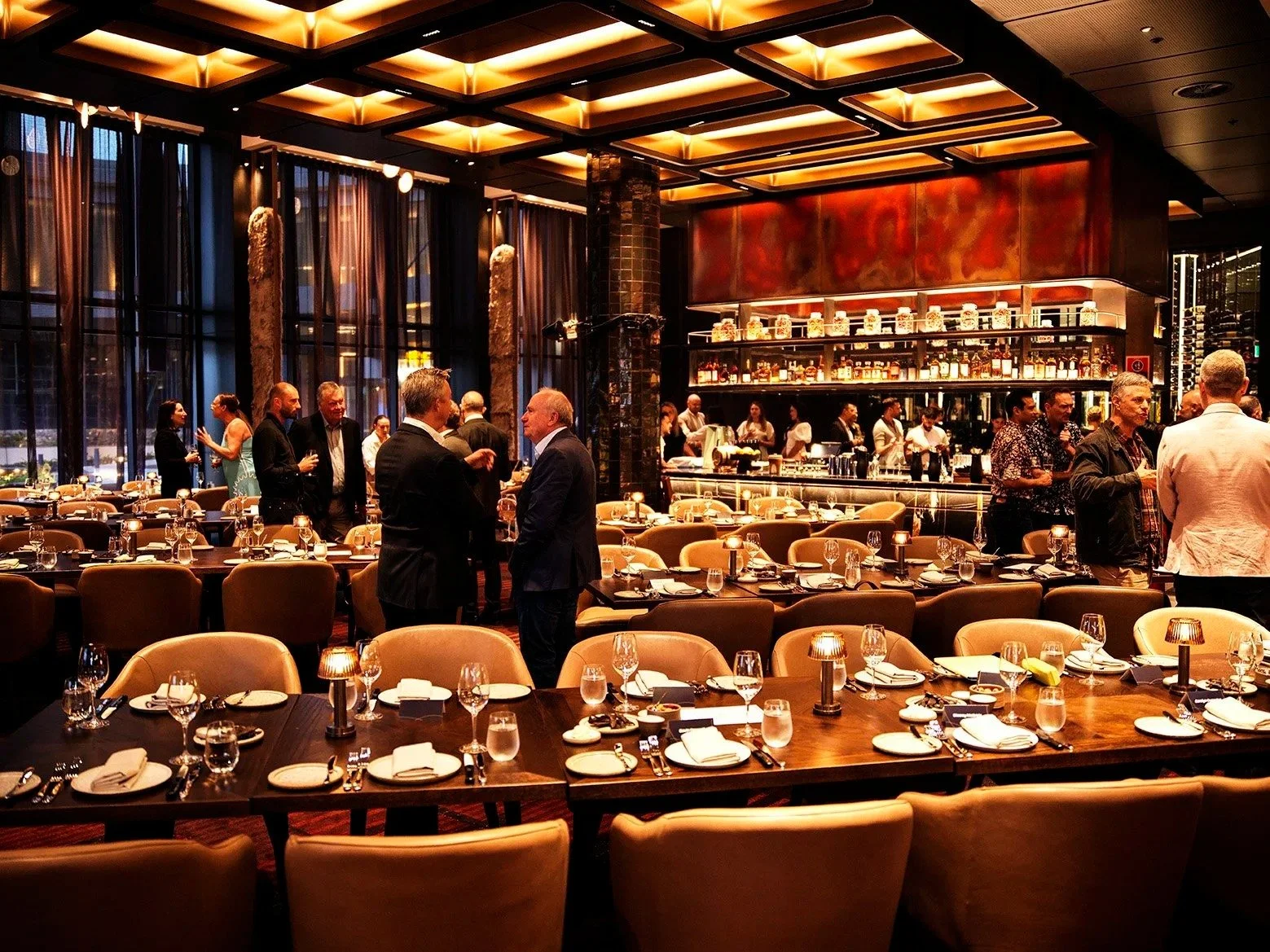 Elegant dinner event in a restaurant with tables set, people mingling, and a well-stocked bar in the background, illuminated with warm lighting.