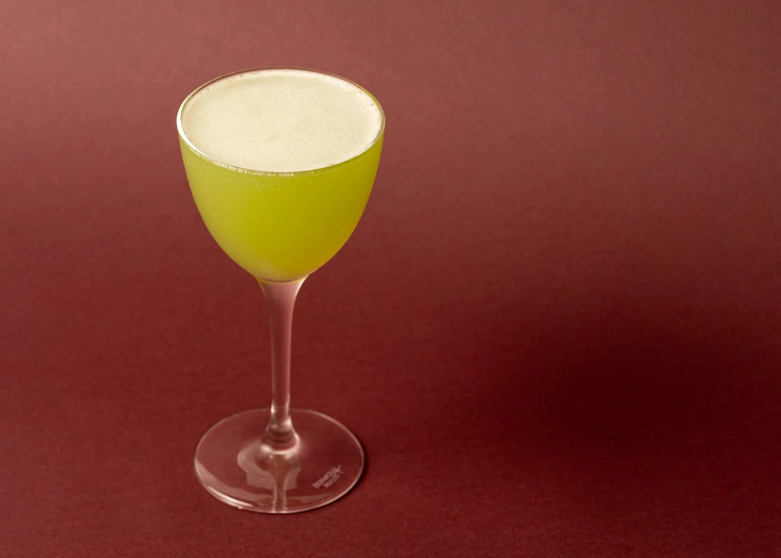 A yellow cocktail or beverage in a clear glass with a thin stem, set against a dark red background.