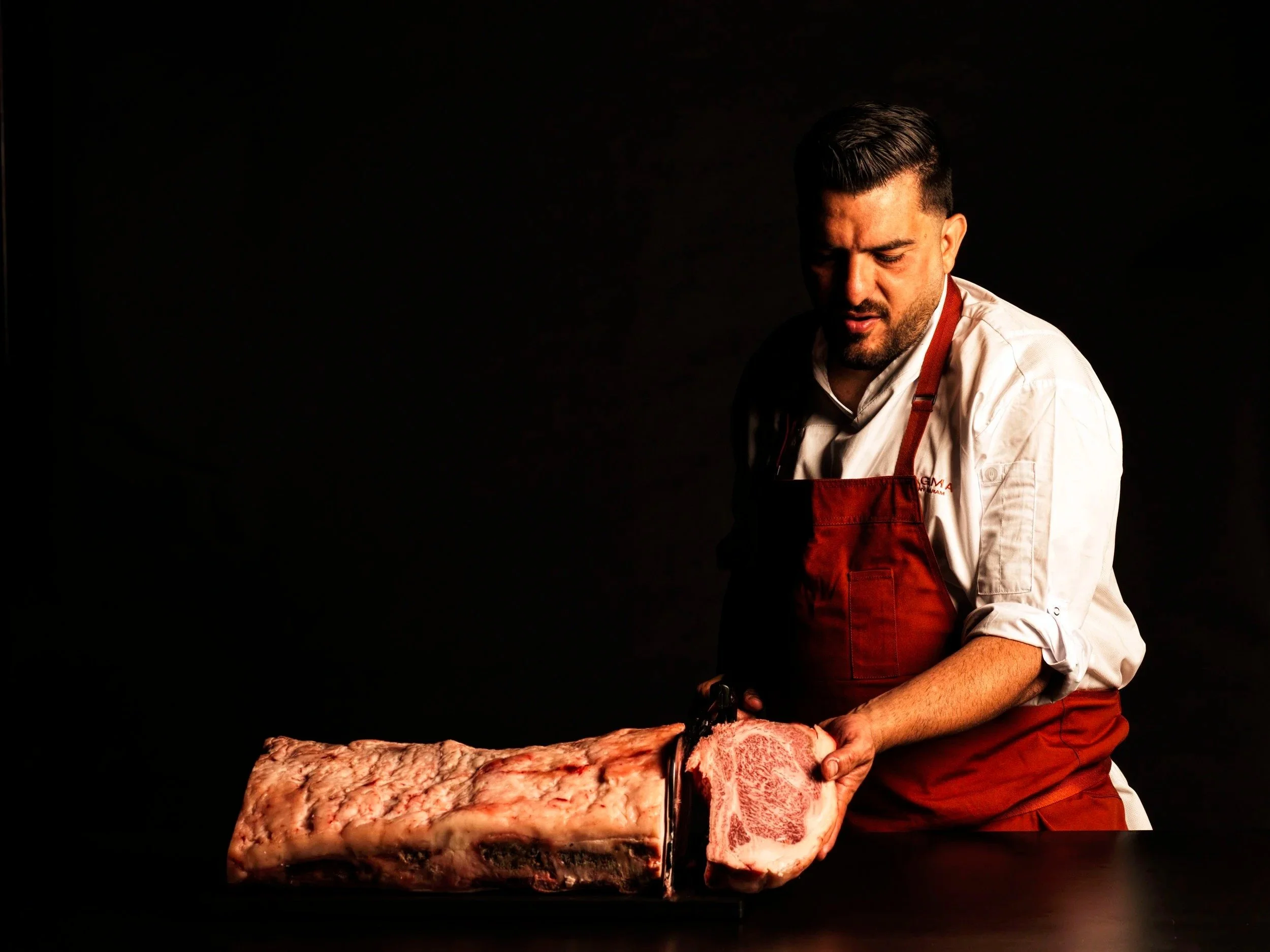 MAGMA BY DANY KARAM sydney. A man wearing a white shirt and red apron is slicing raw meat on a cutting board against a dark background.