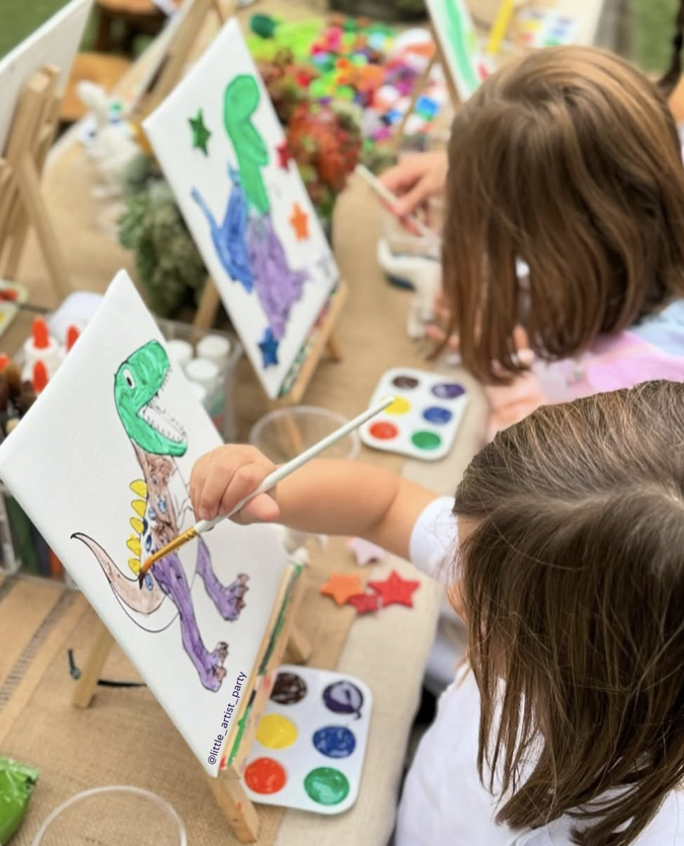 Two children painting colorful dinosaur designs on canvases at an outdoor art table, with paint palettes and star-shaped decorations nearby. kids soiree canvas painting