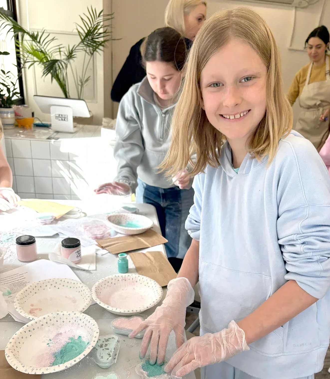 Young girl wearing a white hoodie and gloves working on a craft project with dyed or painted materials on a table, surrounded by bowls and papers, in a room with other children and adults. kids soiree craft activity parties