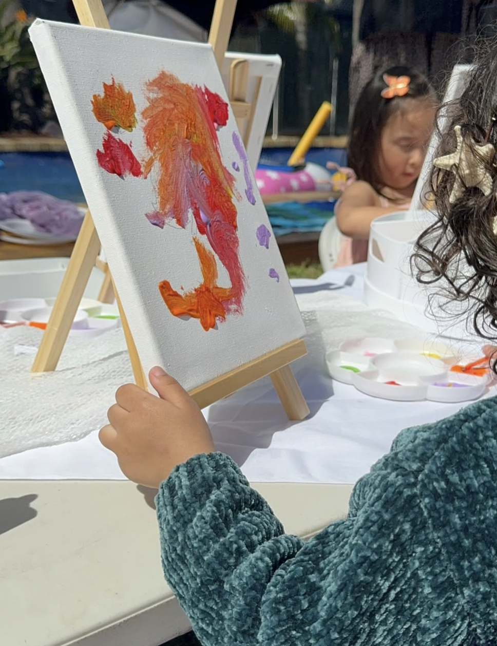 Child painting on a canvas with colorful brushstrokes, with another girl in the background at an outdoor art activity. kids soiree canvas painting party