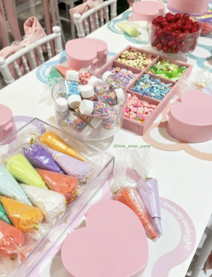 Table decorated for a party with pink and pastel-colored party supplies, including an assortment of colorful candies, sprinkles, cherries in a bowl, and sealed icing bags with pastel-colored frosting.kids soiree jewellery box decoden party