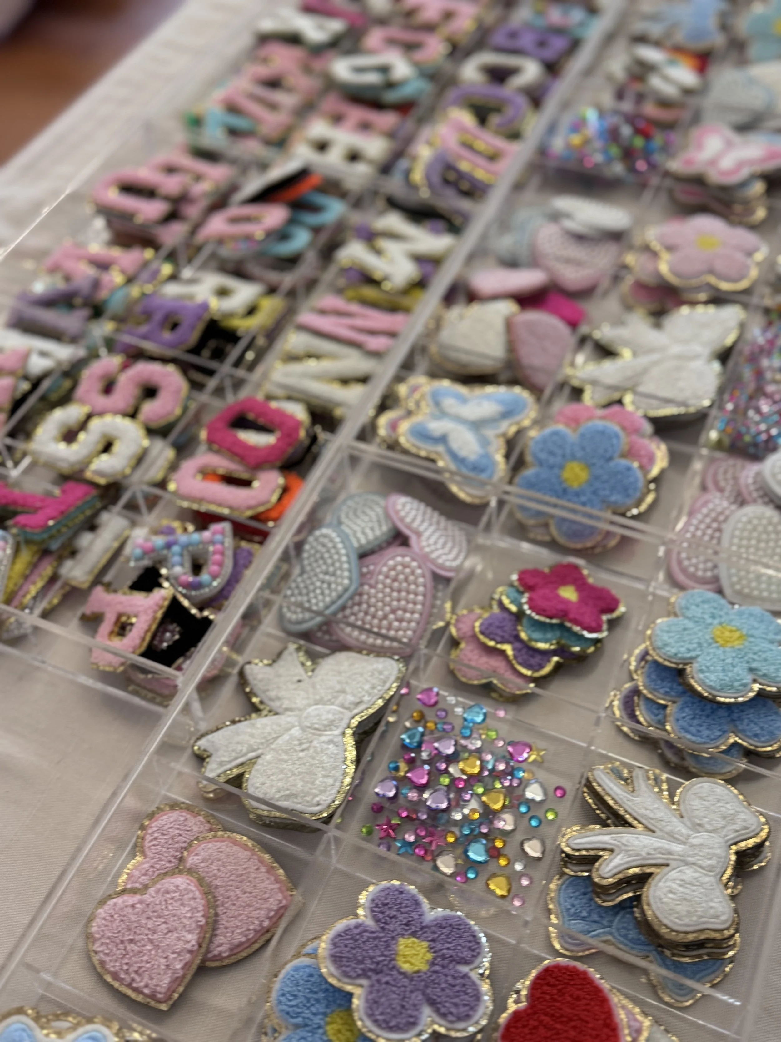 Assorted decorated patches and patches of various shapes and colors, including hearts, flowers, stars, and butterflies, with embellishments like glitter, rhinestones, and embroidery, displayed in clear plastic organizers. kids soiree craft activity