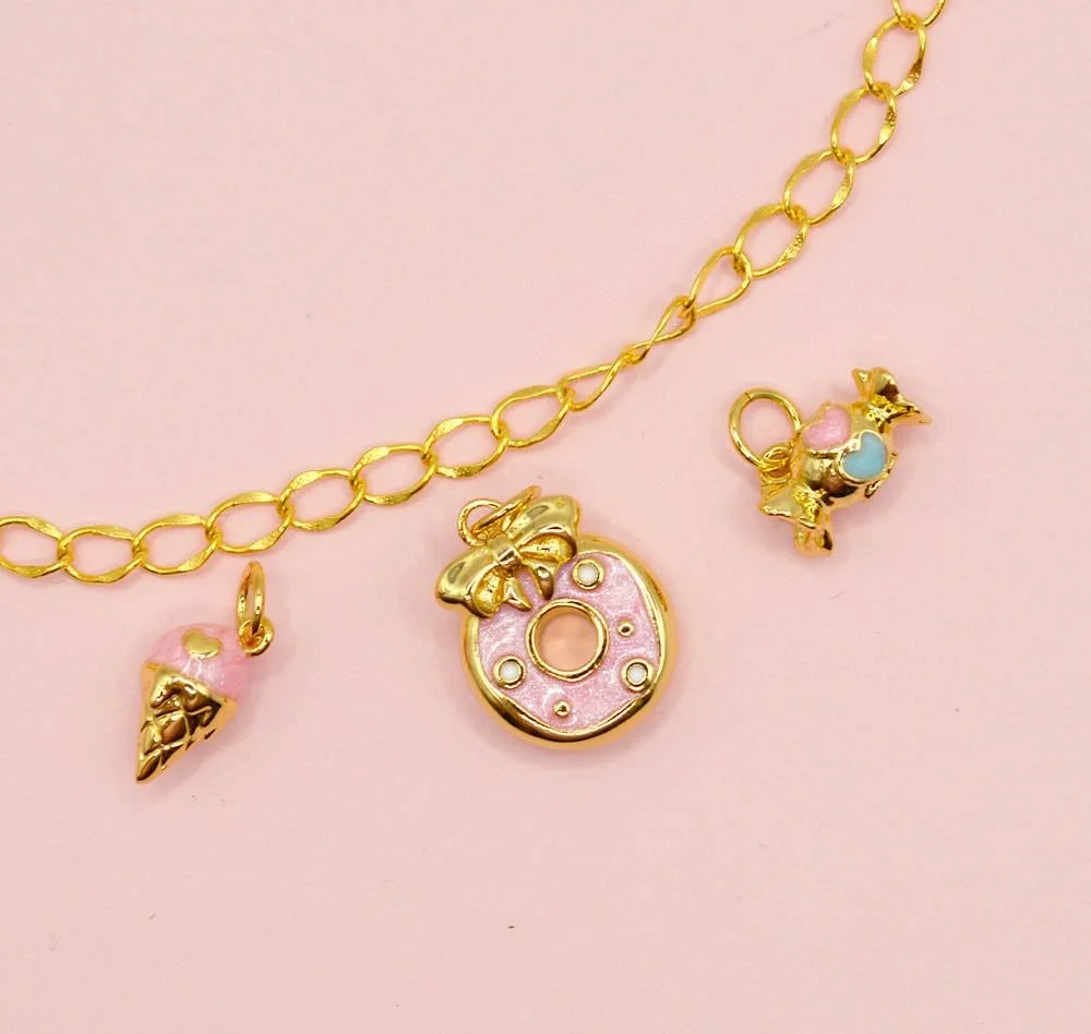 Gold chain bracelet with pink accents and three charms: an ice cream cone, a doughnut with a bow, and a pig with pink and blue patches, all laid on a pink background. kids soiree craft activity parties