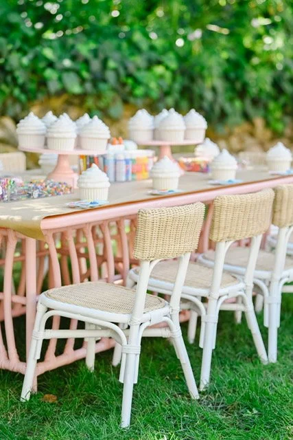 A pastel-colored outdoor cupcake decorating station with cupcakes, frosting, and sprinkles on a pink table, surrounded by wicker chairs and lush green foliage. kids soiree craft activity parties