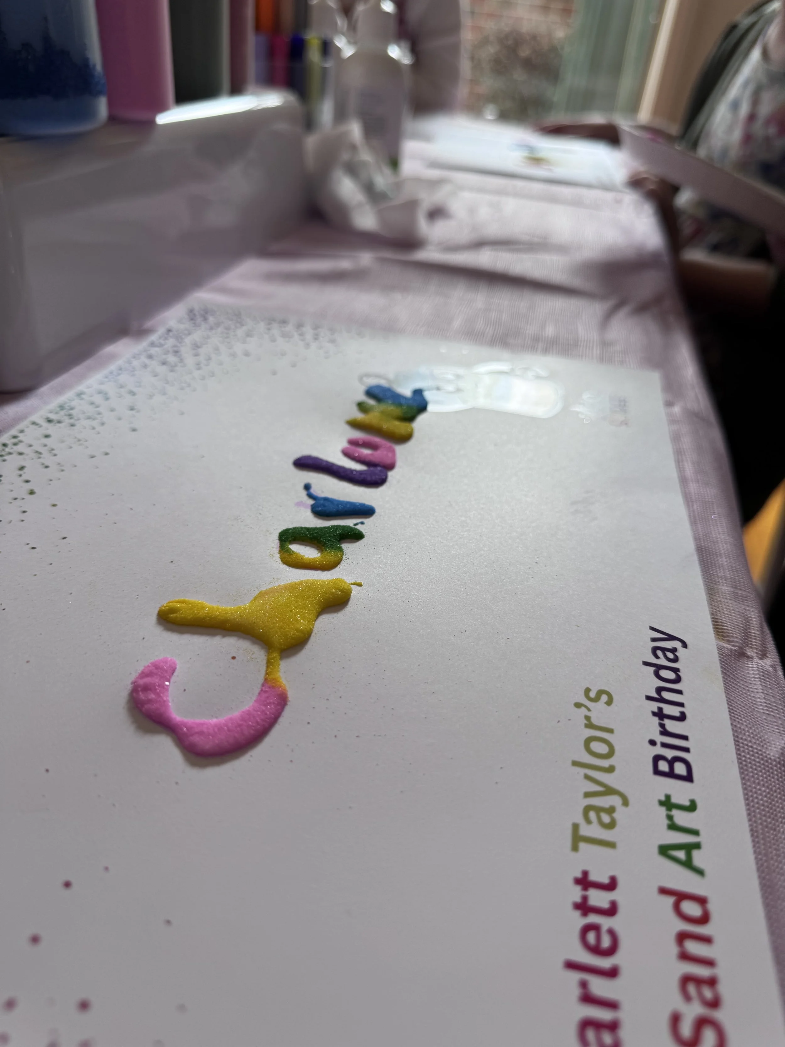 Colorful word spelling 'birthday' in glitter, laid out on a white surface with a partial printed text and a blurred background of art supplies and a window. kids soiree craft activity parties