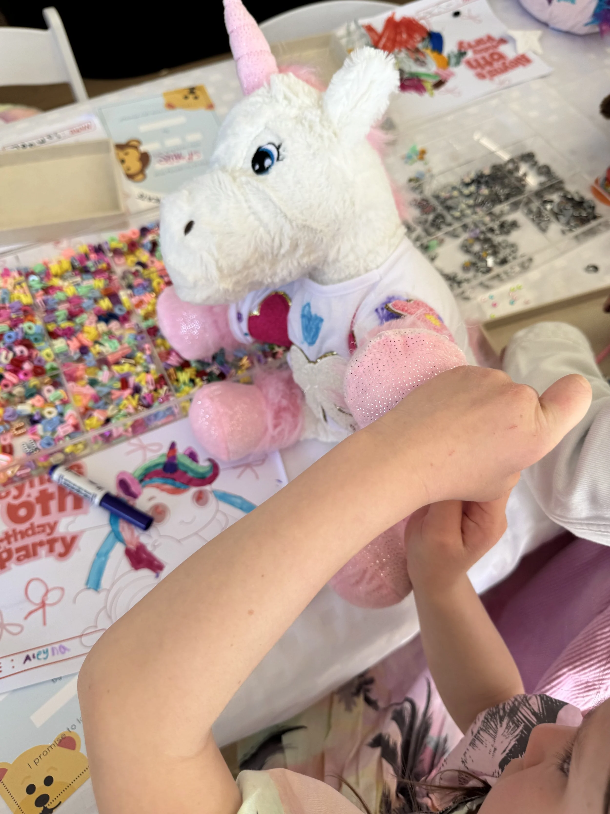 Child holding a plush unicorn toy with a pink horn, white body, and wearing a shirt with a heart on it, surrounded by colorful craft supplies and a birthday party table. kids soiree adopt a plushie party