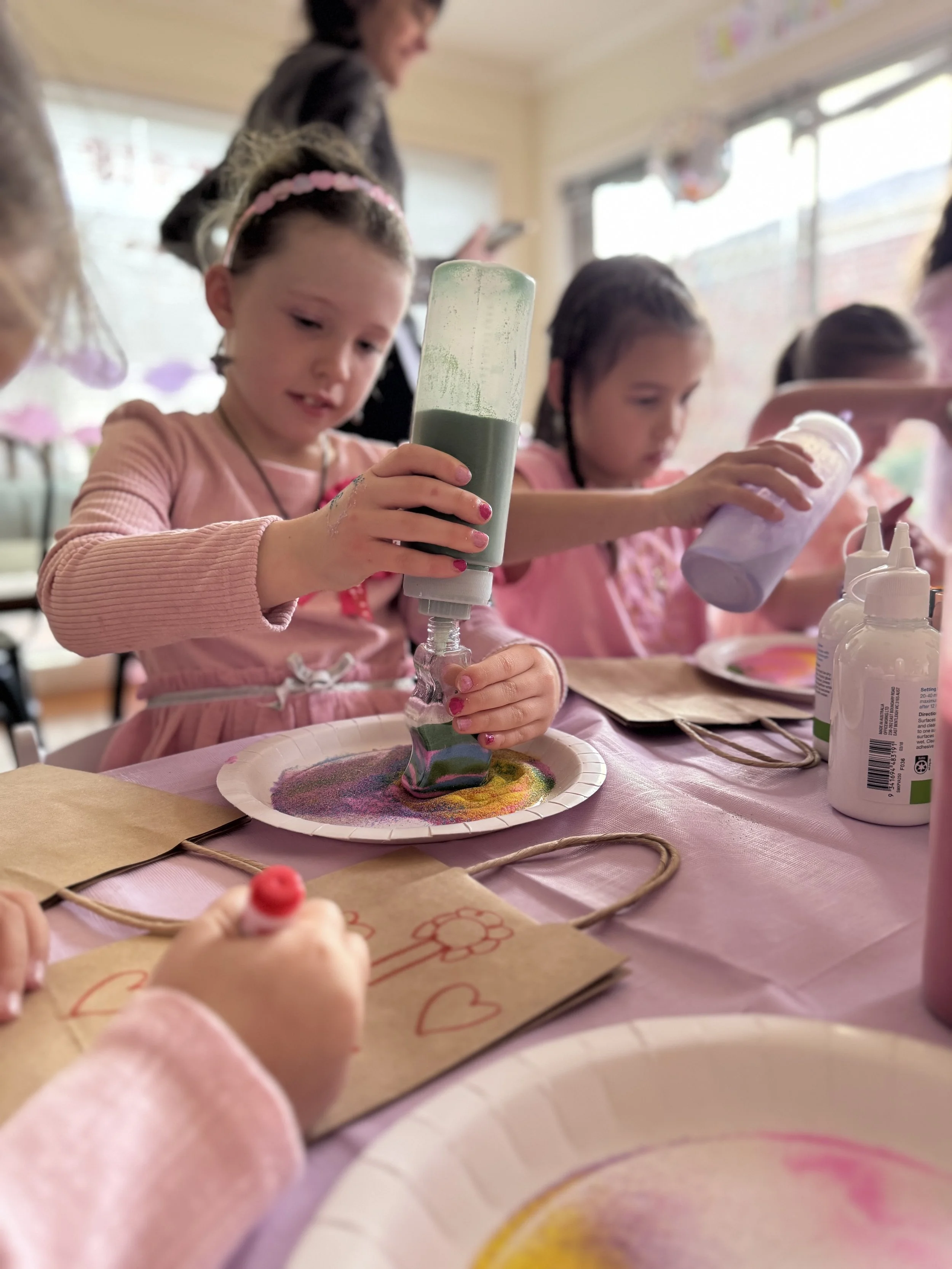 Young children participating in an art activity at a table with colorful sand art bottles, paper bags with heart drawings, and art supplies inside a classroom or craft room. kids soiree craft activity parties