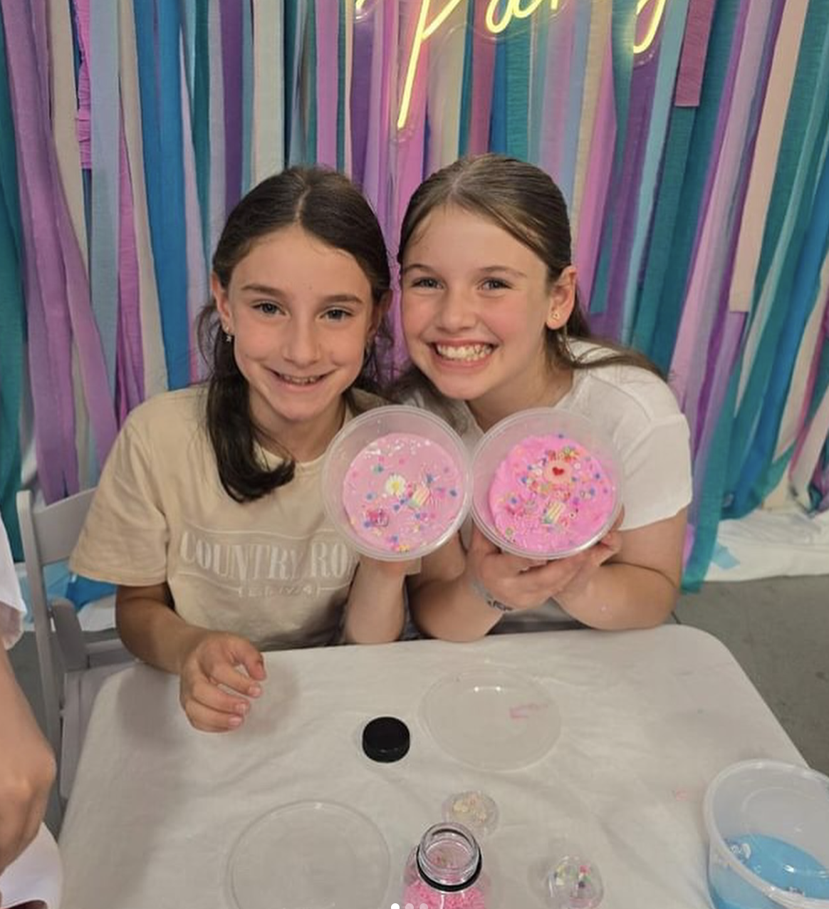 Two young girls smiling and holding containers of pink slime with colorful sprinkles, seated at a table with slime supplies, in front of a pastel-colored ribbon backdrop. Kids Soiree slime party