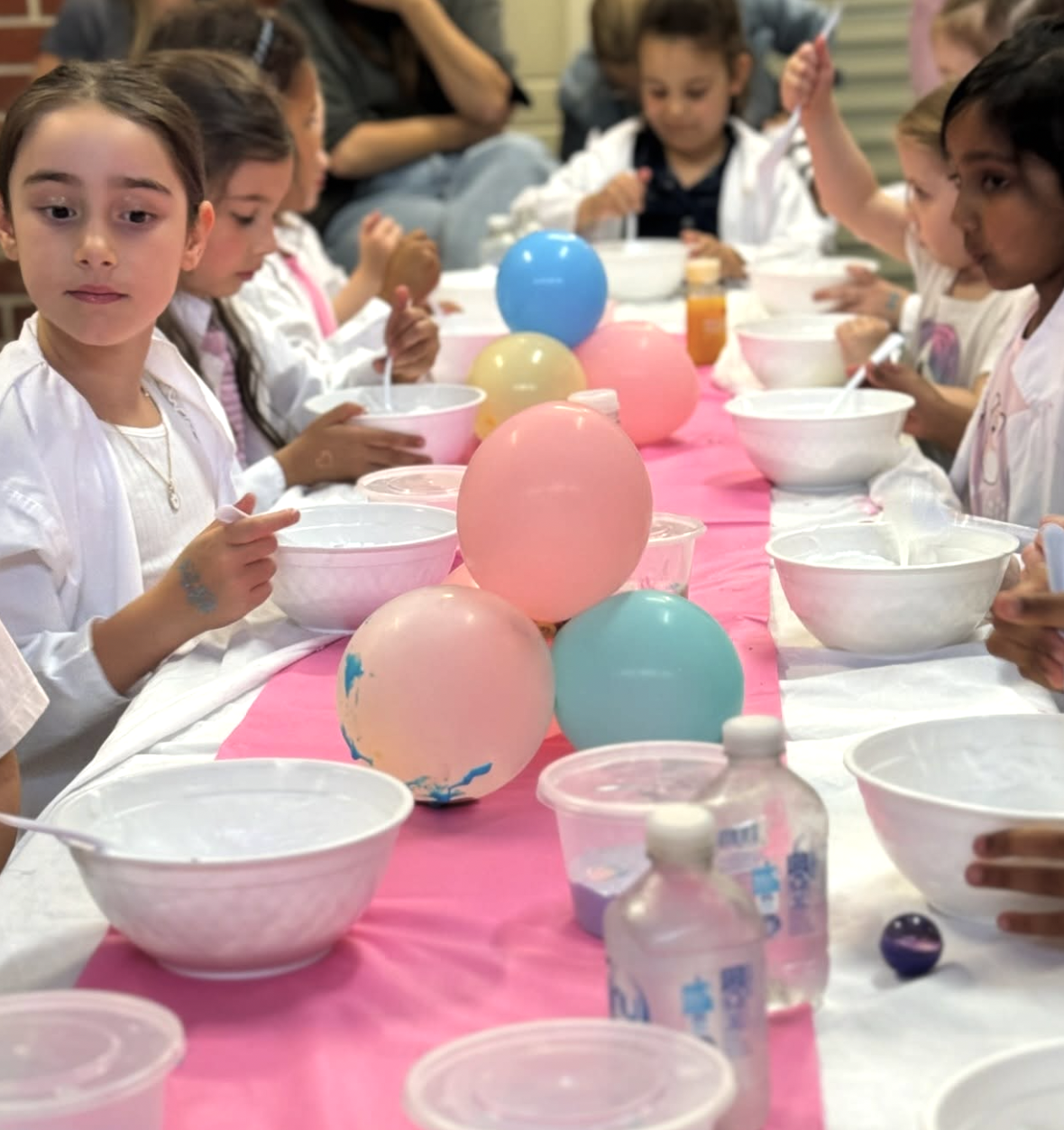 Children sitting at a long table decorated with pink and blue balloons, preparing to eat or do an activity, with bowls and bottles on the table. Kids Soiree Slime Party