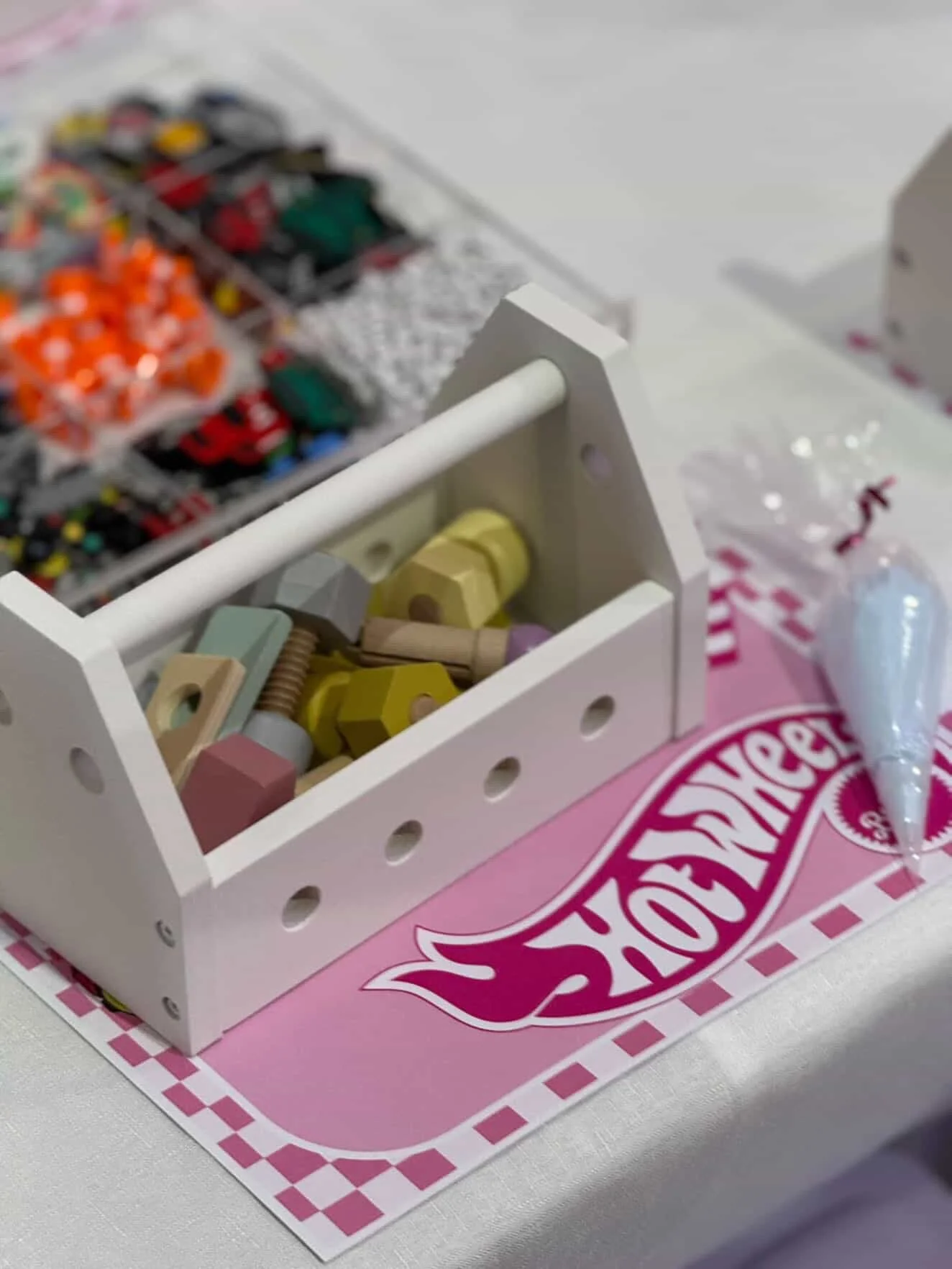 Close-up of a white wooden toy chest filled with colorful toy hammer blocks, placed on a pink gingham-patterned tablecloth. Blurred various small toys are visible in the background. kids soiree craft activity parties