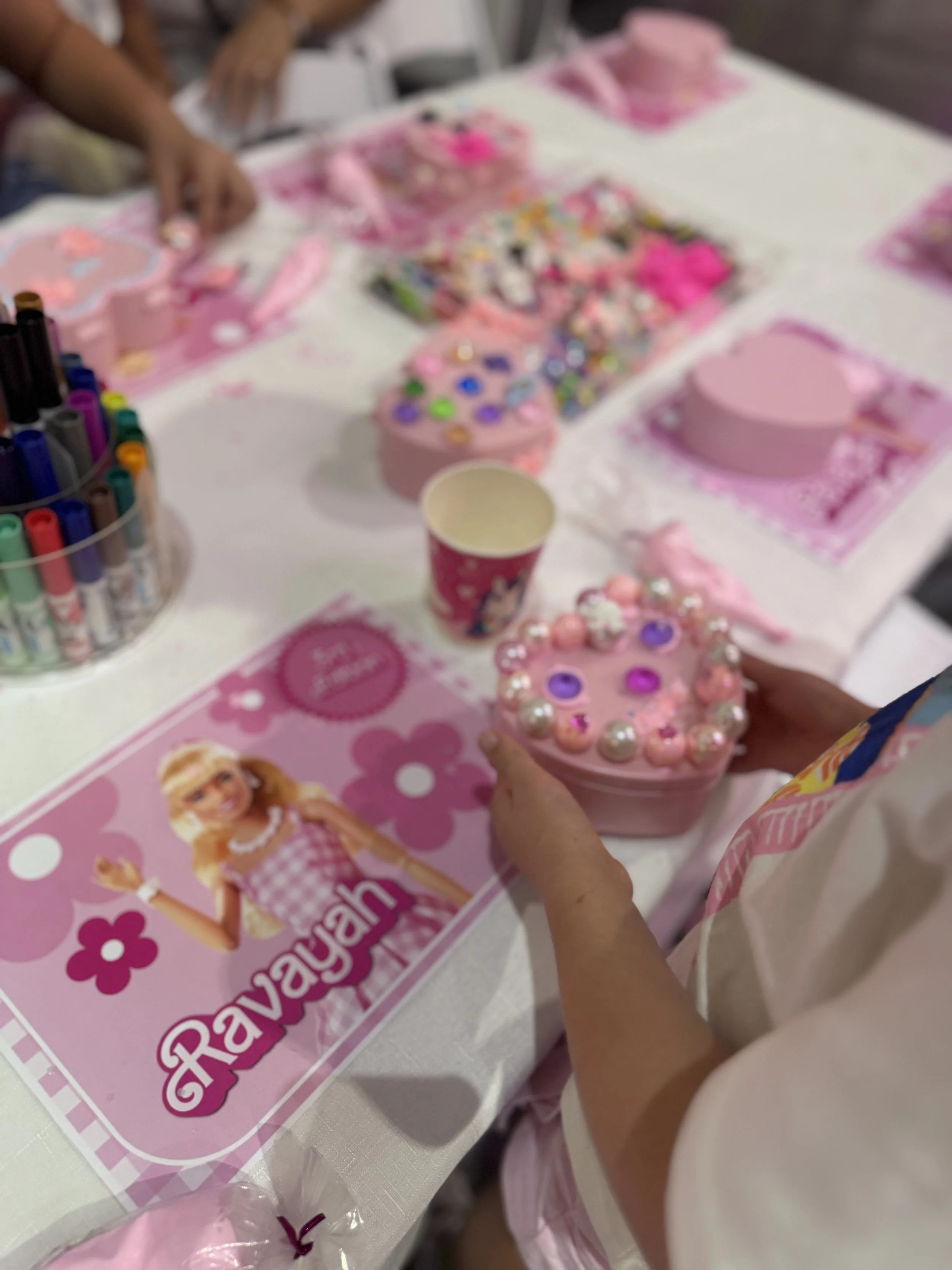 A person holding a pink cupcake decorated with multi-colored pearls and pink frosting at a table with a pink tray of similar cupcakes, a flyer with a doll illustration, and various art supplies.kids soiree jewellery box decoden party