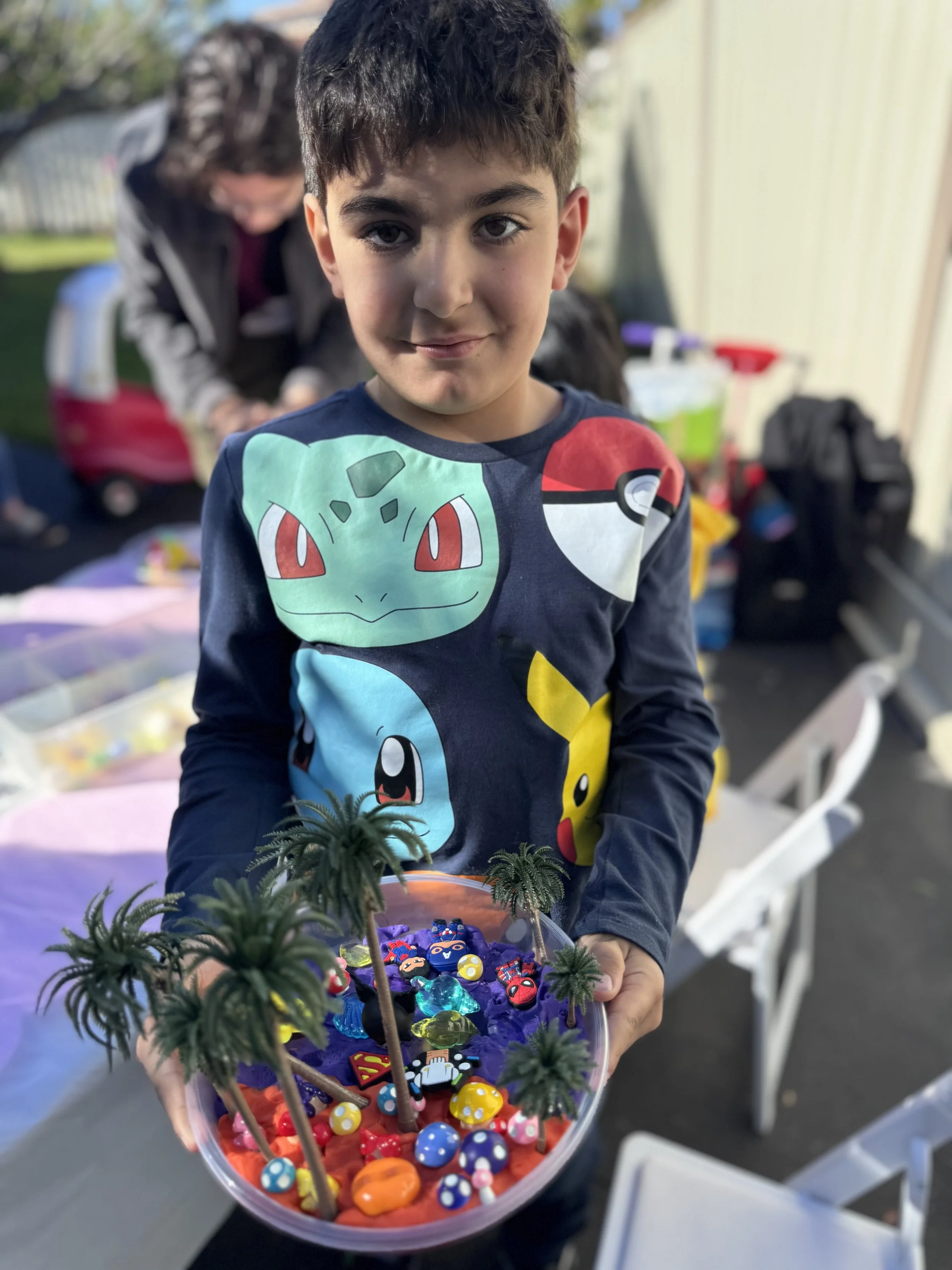 A young boy holding a clear plastic bowl filled with colorful small toys, picture stones, and decorative items, with miniature palm trees made of wire and beads inside the bowl. kids soiree craft activity parties