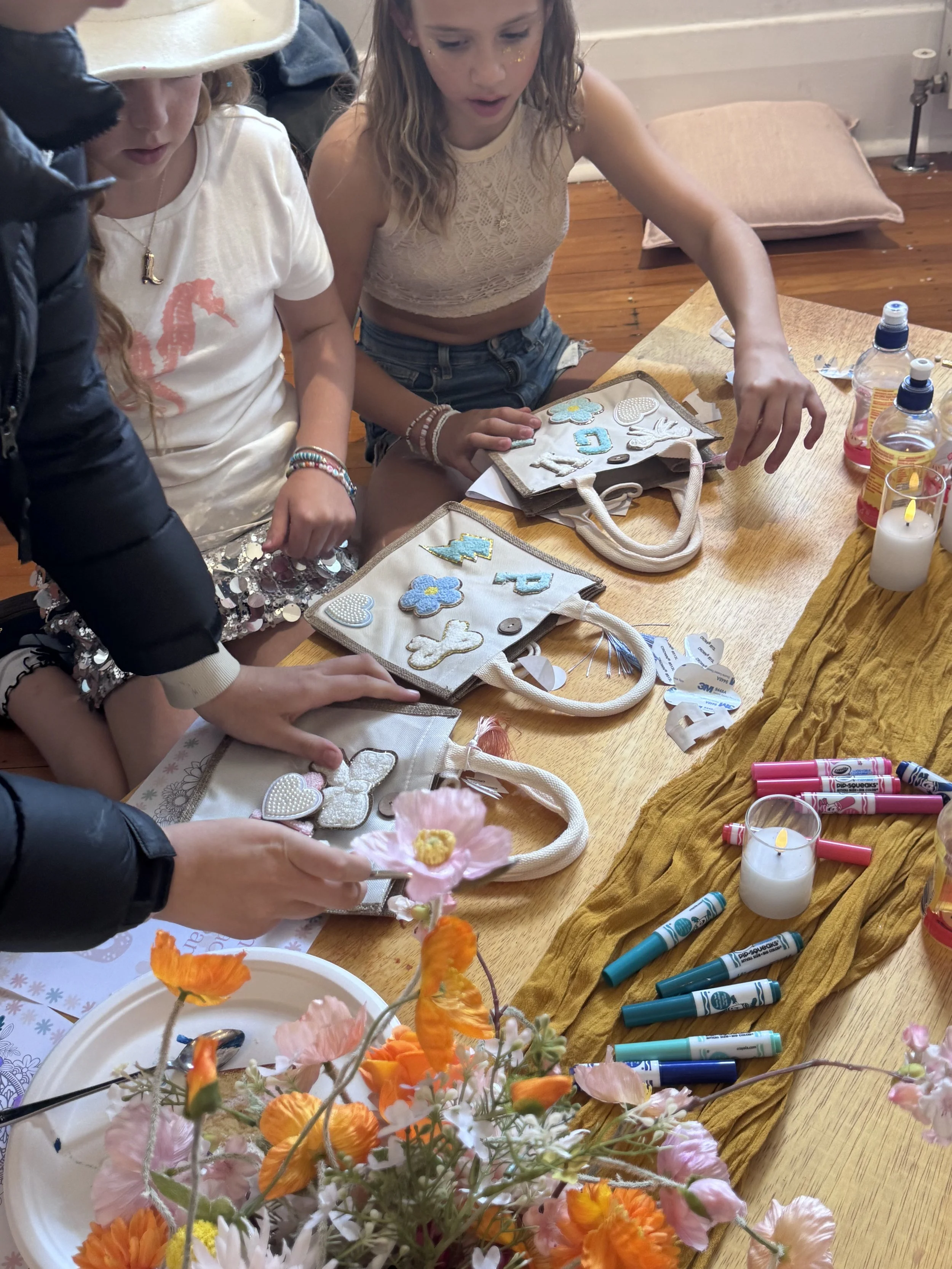 Children looking at decorated patches and craft supplies on a table, with flowers in the foreground, during a craft activity. kids soiree craft activity parties