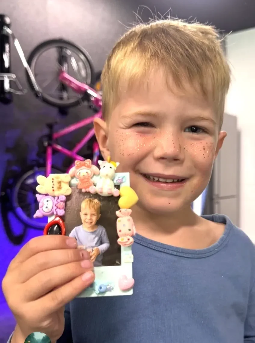 A young boy with red hair, freckles, and a blue shirt is smiling and winking at the camera. He is holding a decorated photograph frame surrounded by colorful toys and figurines. In the background, kids soiree craft activity parties