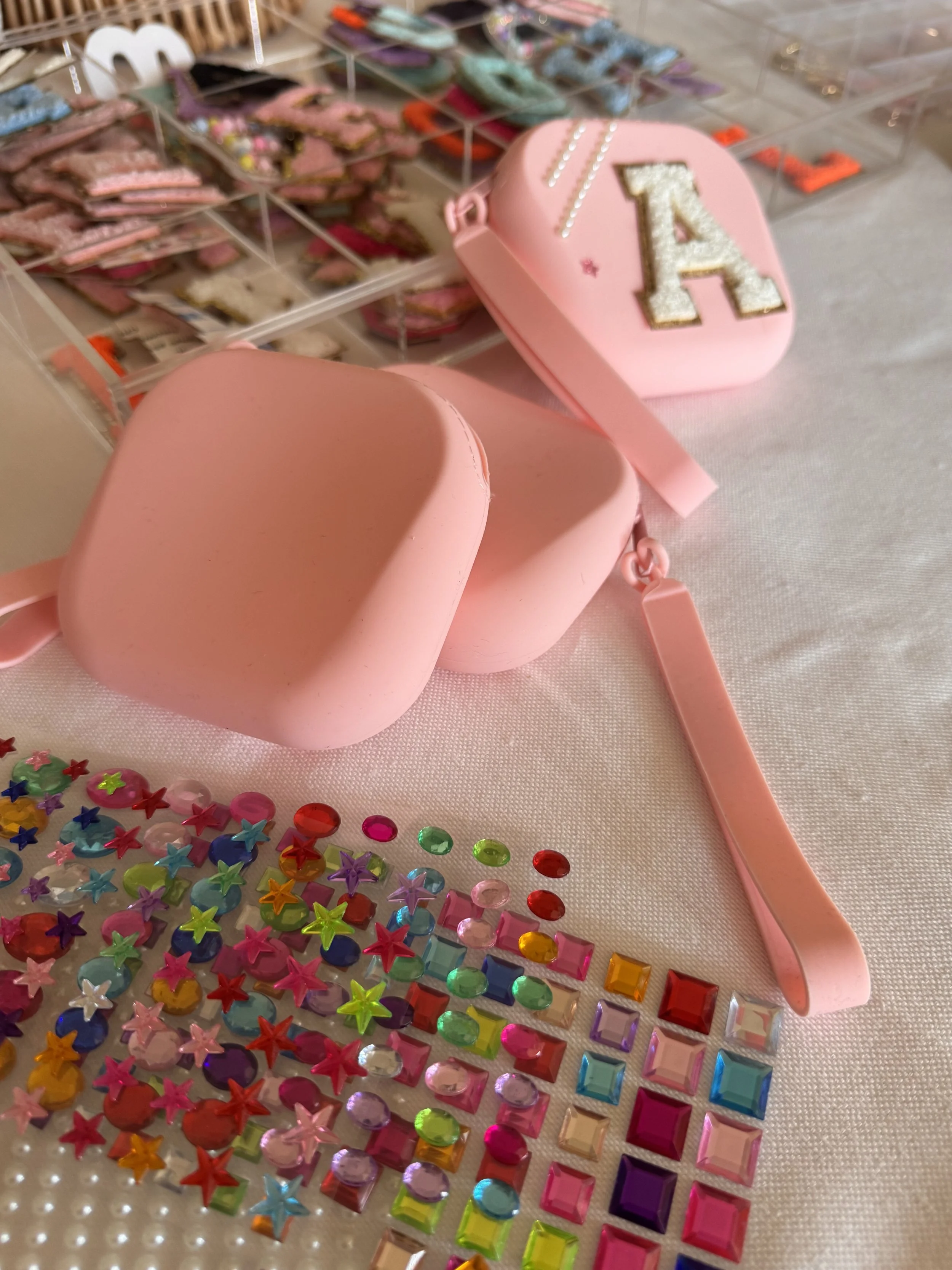 Display of colorful decorative rhinestones, sequins, and a pink makeup bag with the letter A on it, set on a white surface. lip gloss making kids soiree