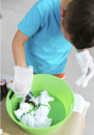 Child wearing gloves and a blue shirt, holding a spray bottle over a green basin filled with white cloths and green liquid. kids soiree craft activity parties