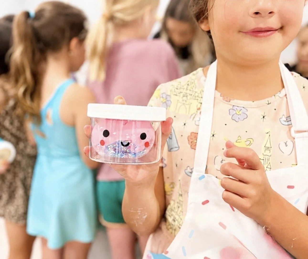 Child holding a clear container with a pink slime or gel inside, decorated with a smiling face, at a group activity or class. kids soiree craft activity parties