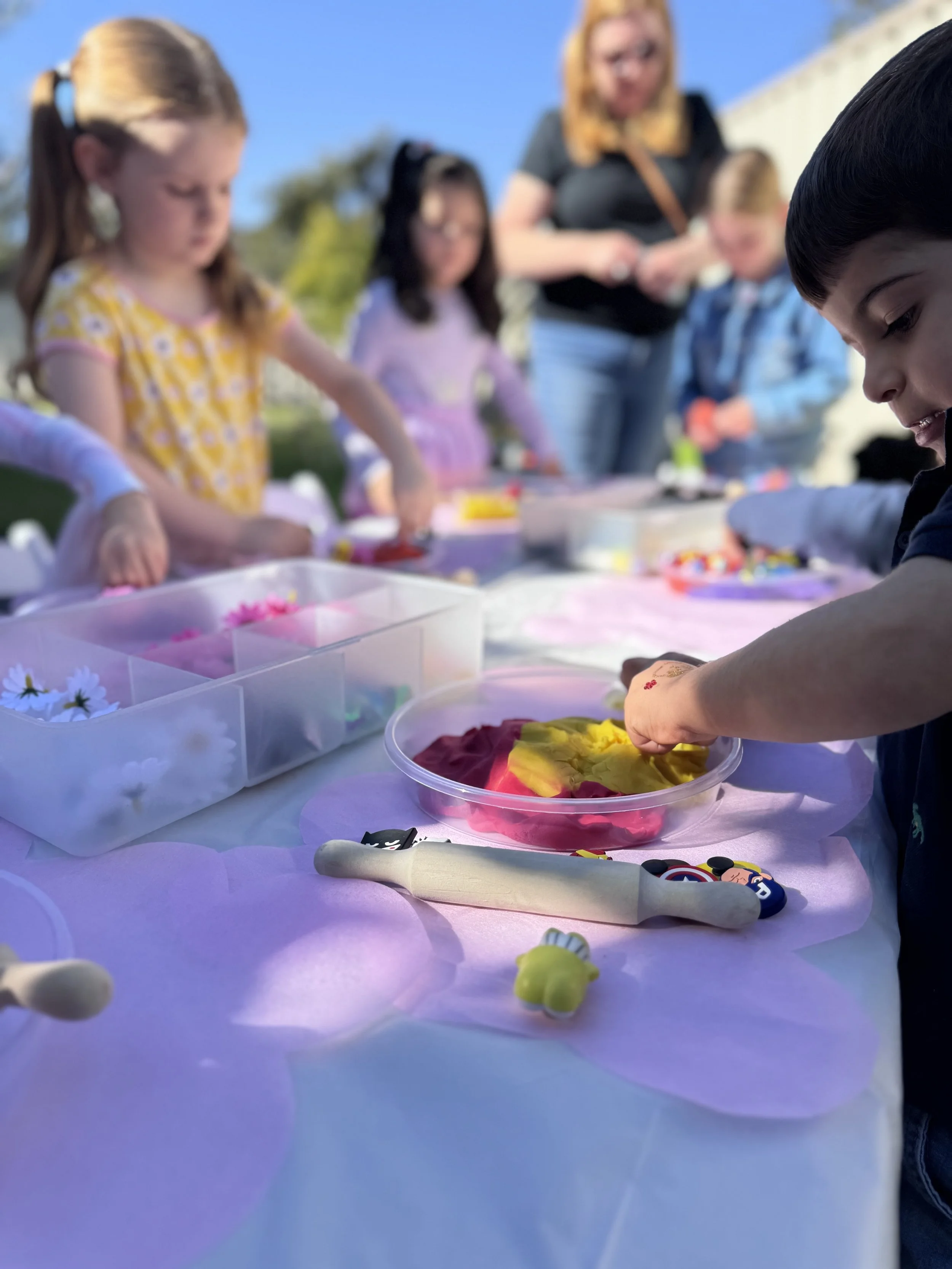 Children and an adult participating in an outdoor craft activity at a table with colorful paper, flowers, and craft supplies. kids soiree craft activity parties