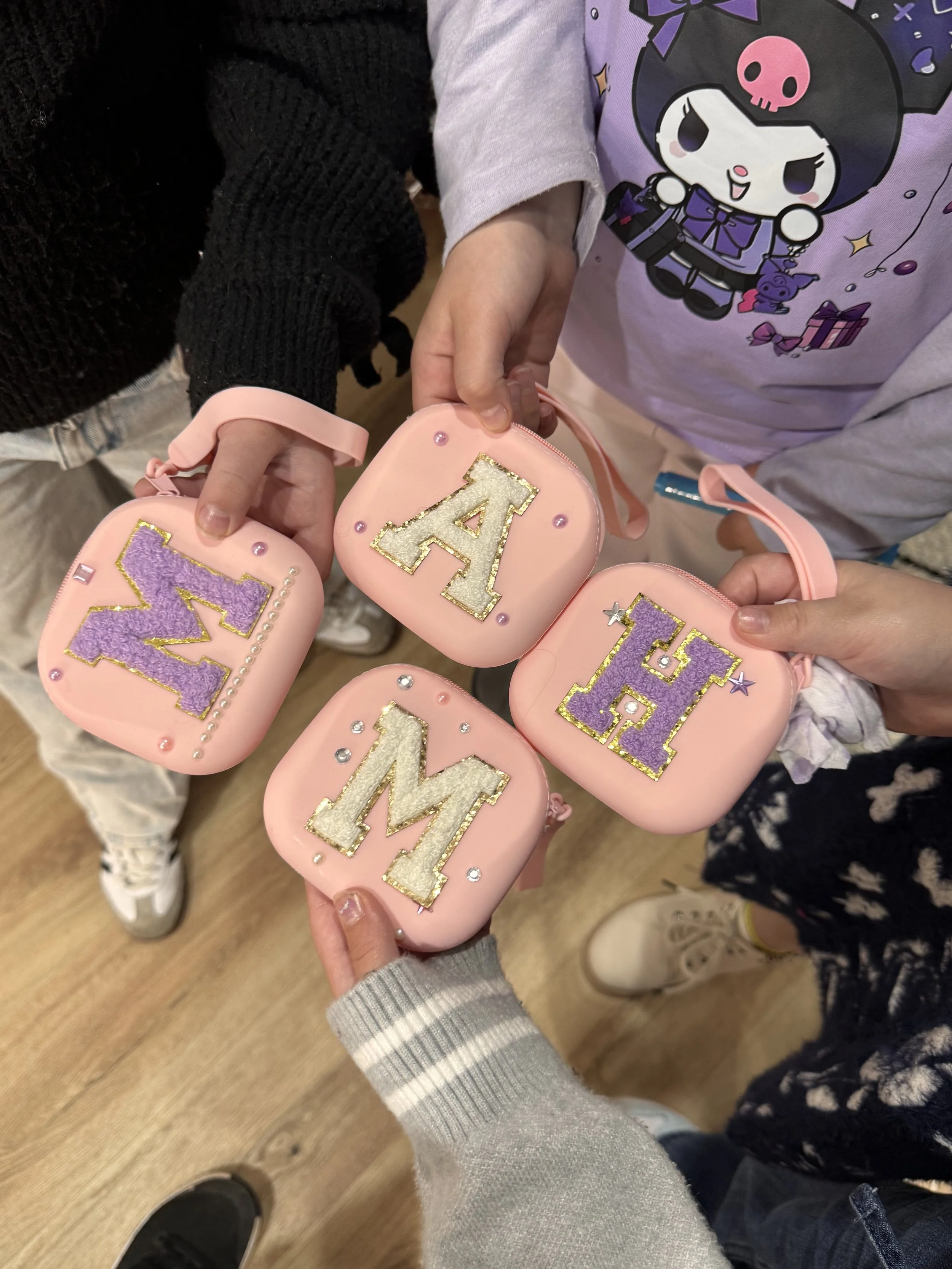 A group of people holding small pink bags with purple, white, and gold glittery letters that spell 'A' and 'M'. The people are wearing casual clothing, including a light purple shirt with a cartoon girl print lip gloss making kids soiree