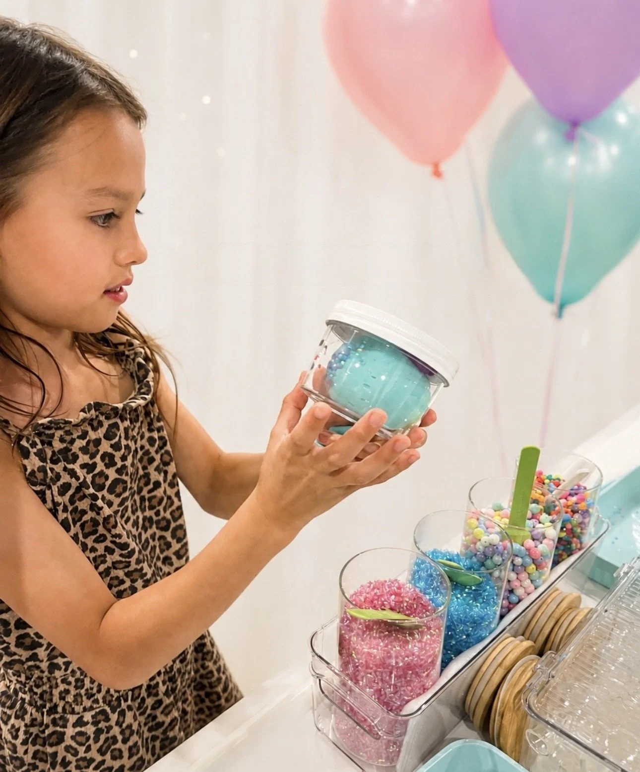 A young girl in a leopard print dress is holding a clear plastic container with blue beads or sprinkles, looking at it. Behind her are pink, purple, and light blue balloons kids soiree craft activity parties