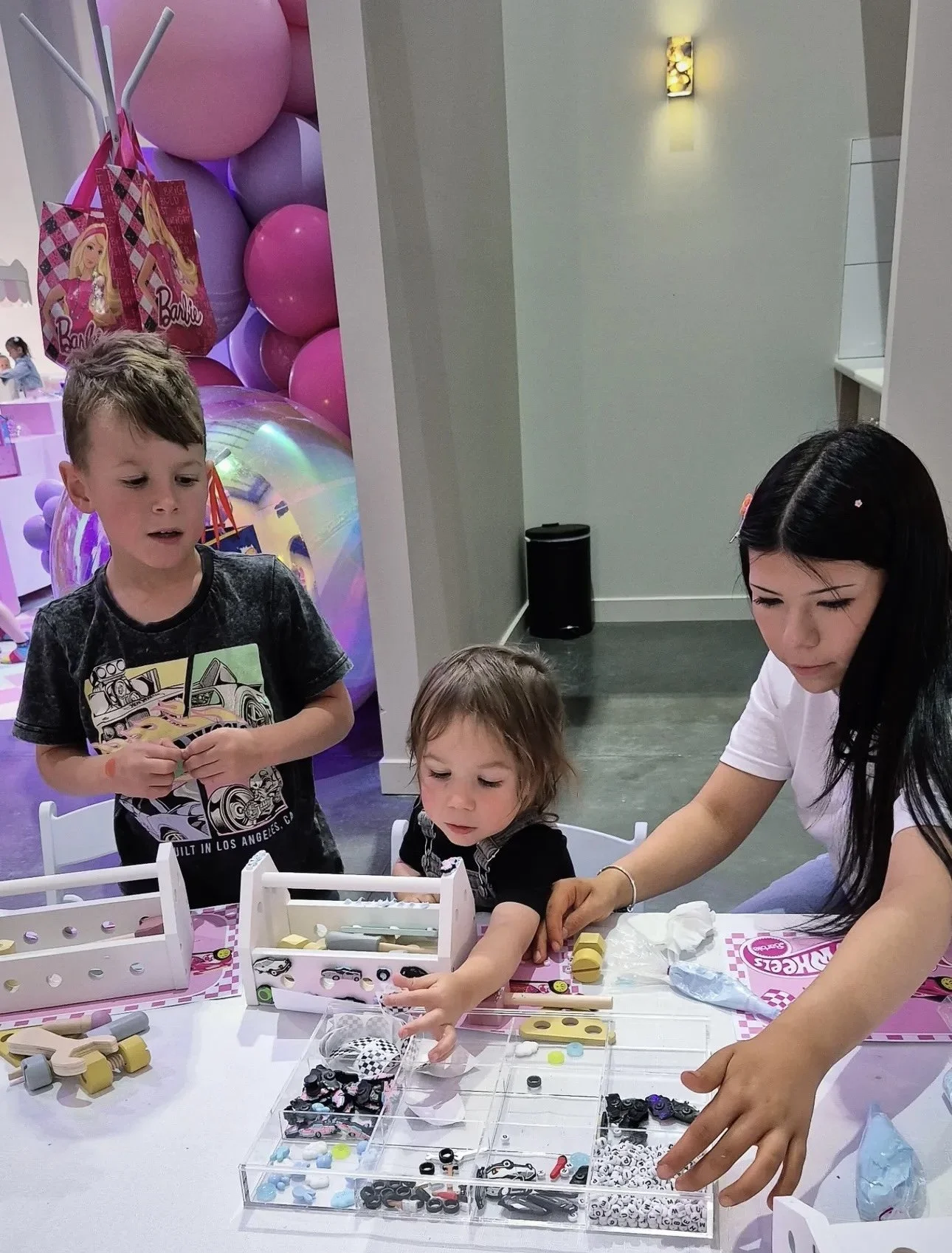 Children and a woman play with small toy cars and accessories at a table in a store or party setting with balloons and pink decorations in the background. kids soiree craft activity parties