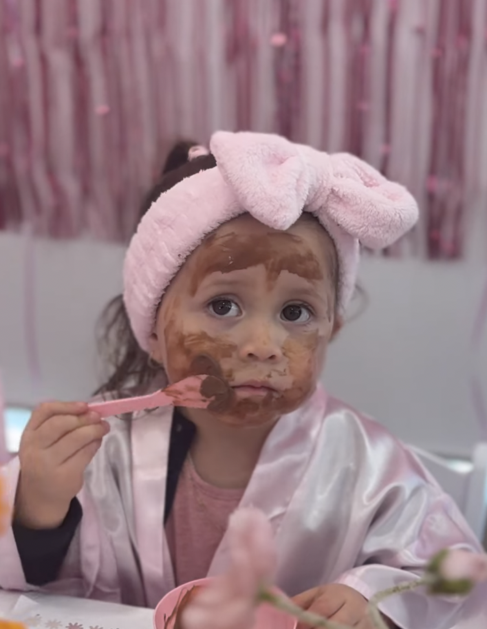 Young girl with brown hair, pink headband with a bow, wearing a pink satin robe, covered in chocolate face mask, holding a pink spoon with chocolate, sitting at a table with a pink backdrop. Kids Soiree pamper party
