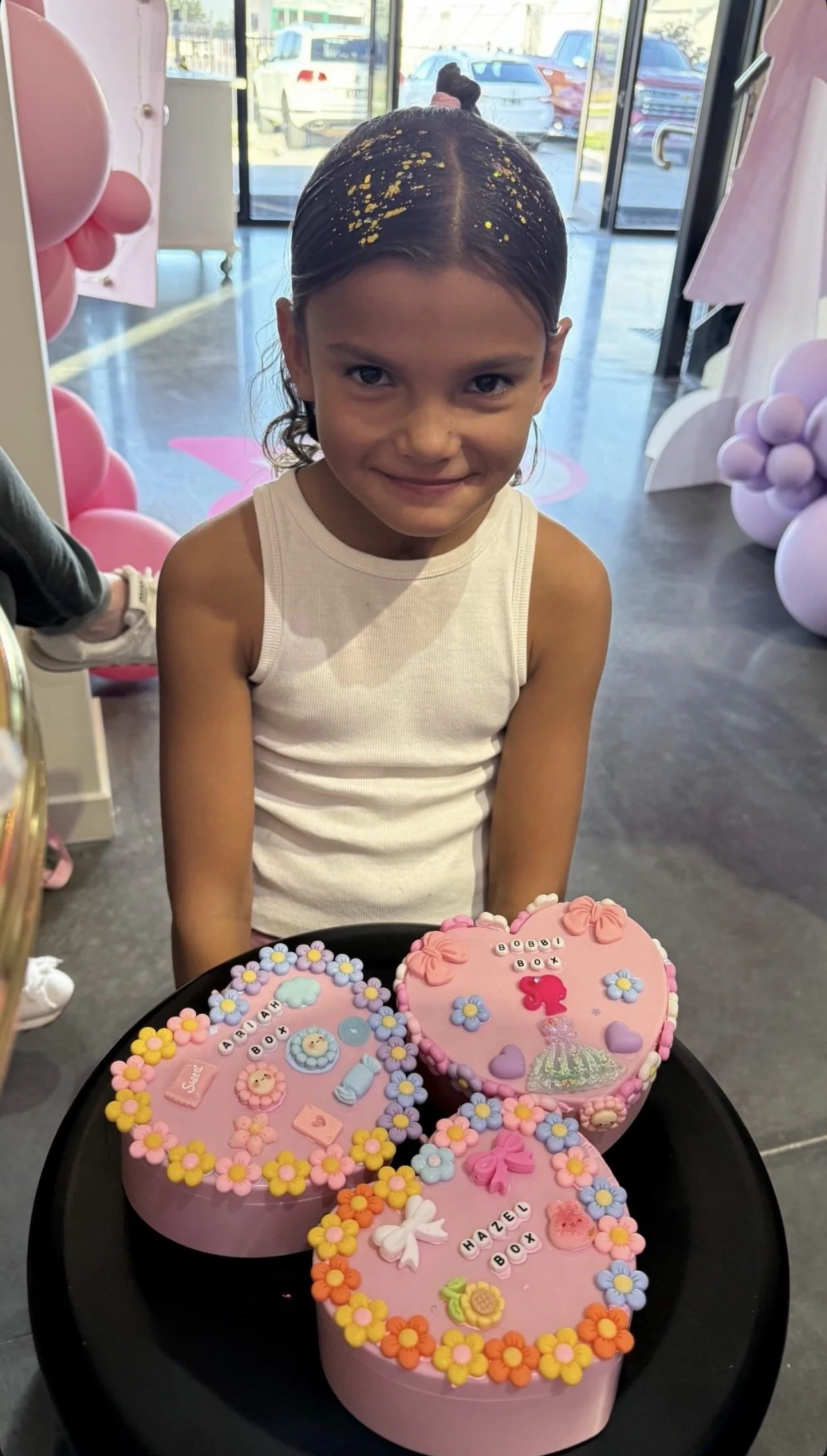 Smiling young girl with dark hair styled in a bun, wearing a white tank top, standing behind a tray of pink decorated cookies and cakes, with a birthday celebration setting featuring pink and purple balloons in the background. kids soiree craft activ