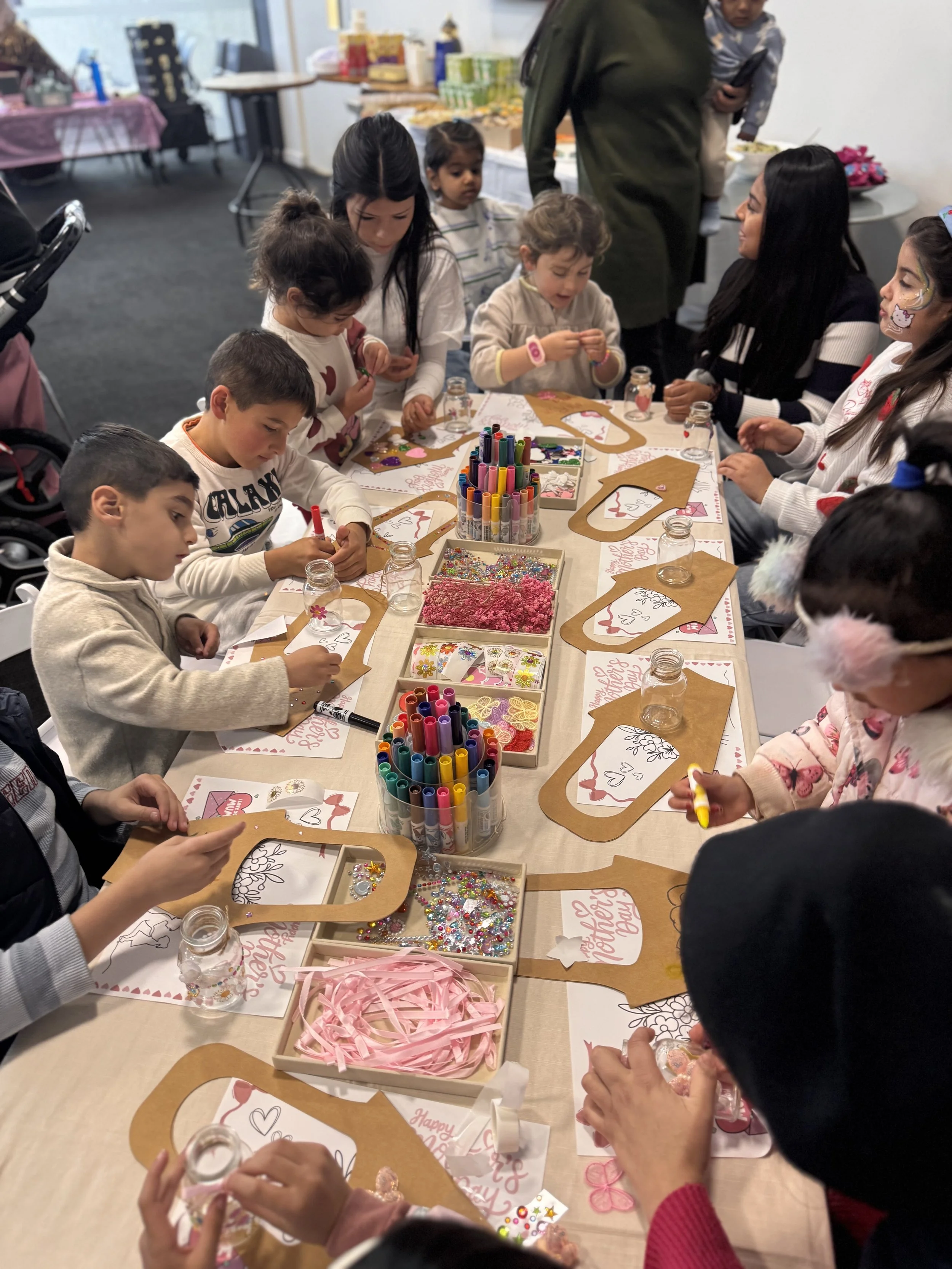 Children and adults gathered around a table making Valentine's Day crafts with markers, glitter, and stickers, with Valentine's Day greeting cards on the table. kids soiree craft activity parties