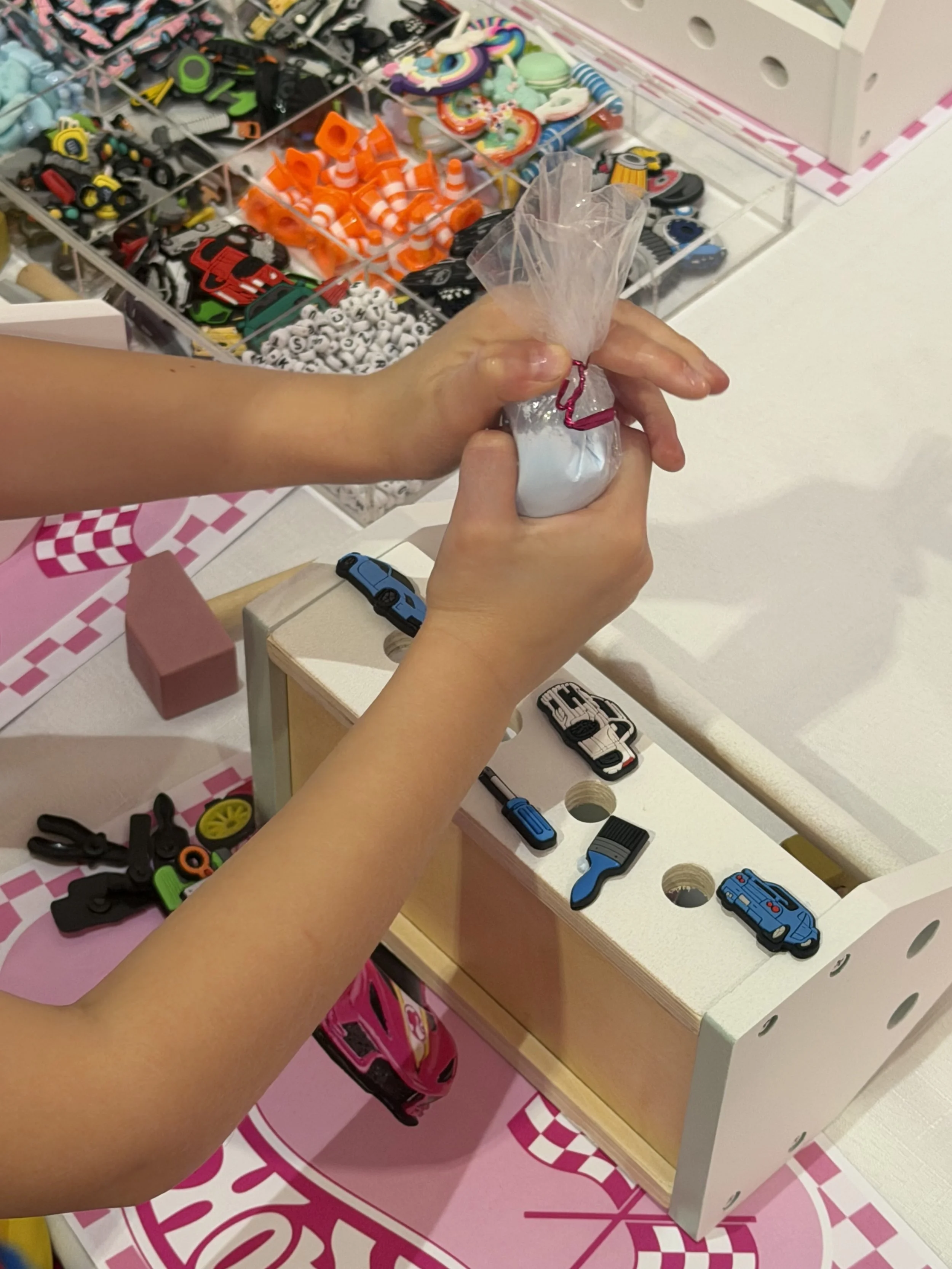 Close-up of a child's hands holding a small bag of white liquid, at a display of toy car accessories and toy vehicles on a table with a pink and white checkered mat. kids soiree craft activity parties