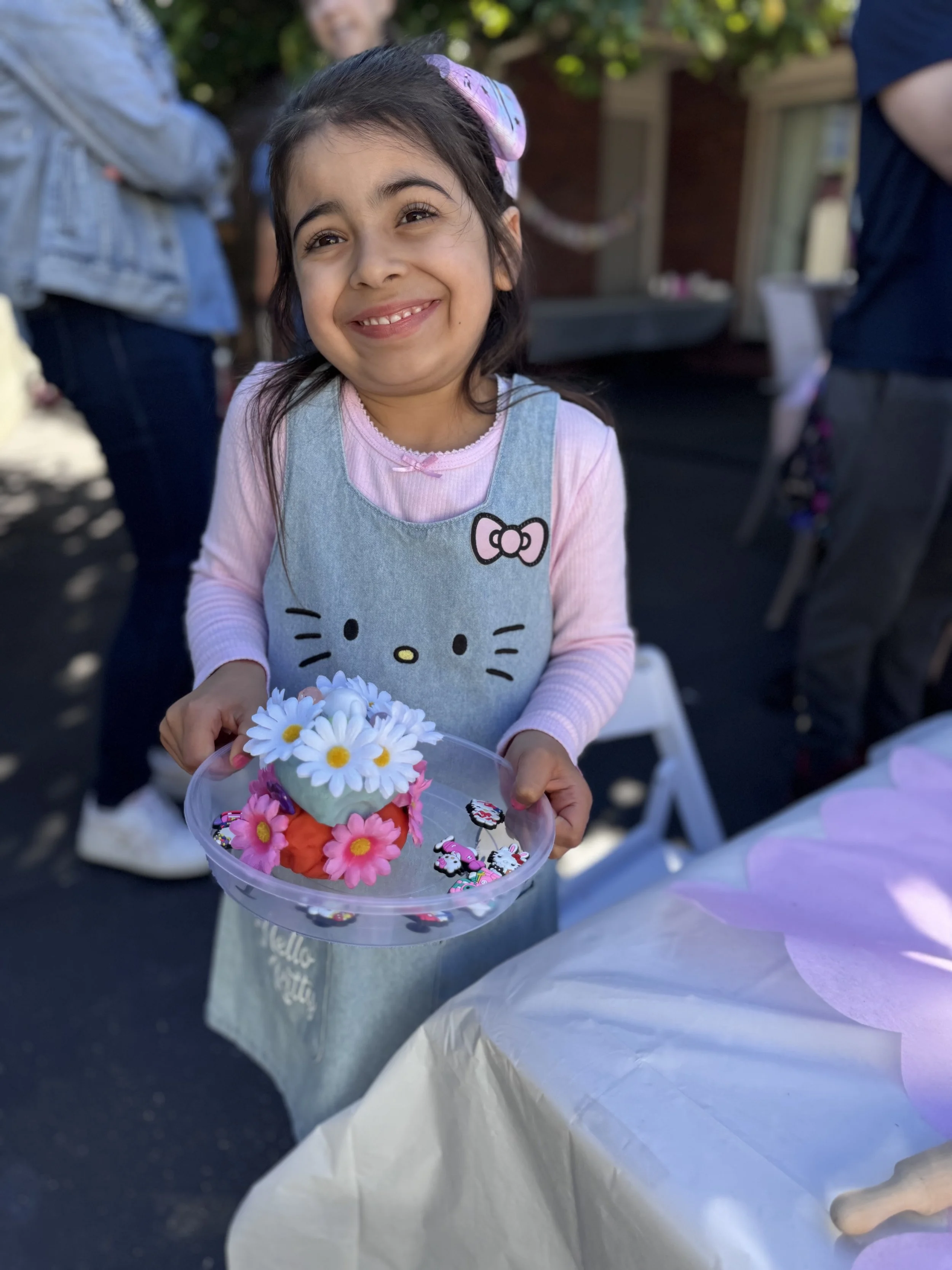 A young girl smiling and holding a clear plastic container filled with colorful flower-shaped accessories and jewelry, outdoors during daytime with people around. kids soiree craft activity parties