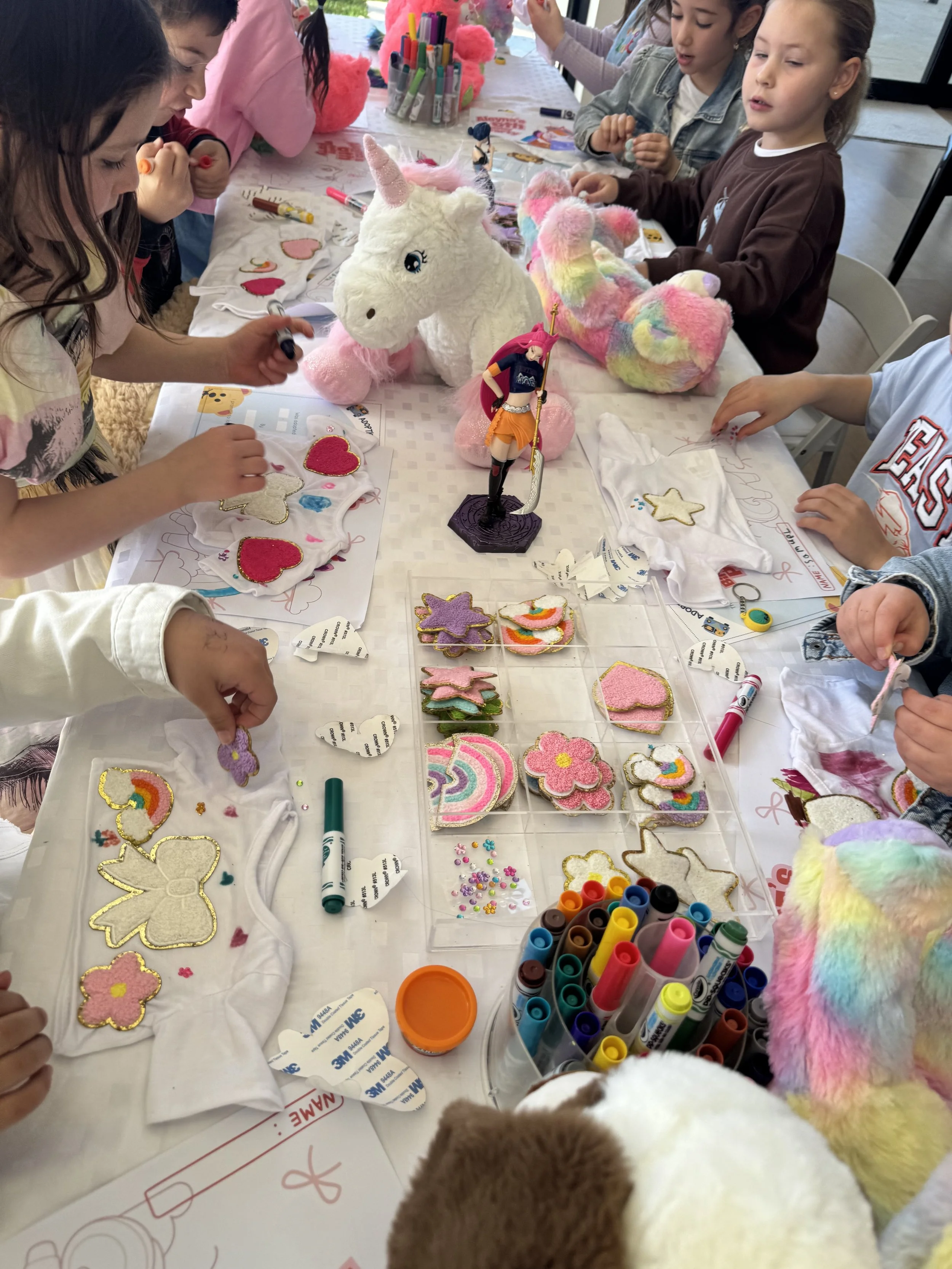 Children participating in a craft activity at a table, surrounded by plush unicorns, decorated felt shapes, markers, and supplies. kids soiree kids birthday craft parties
