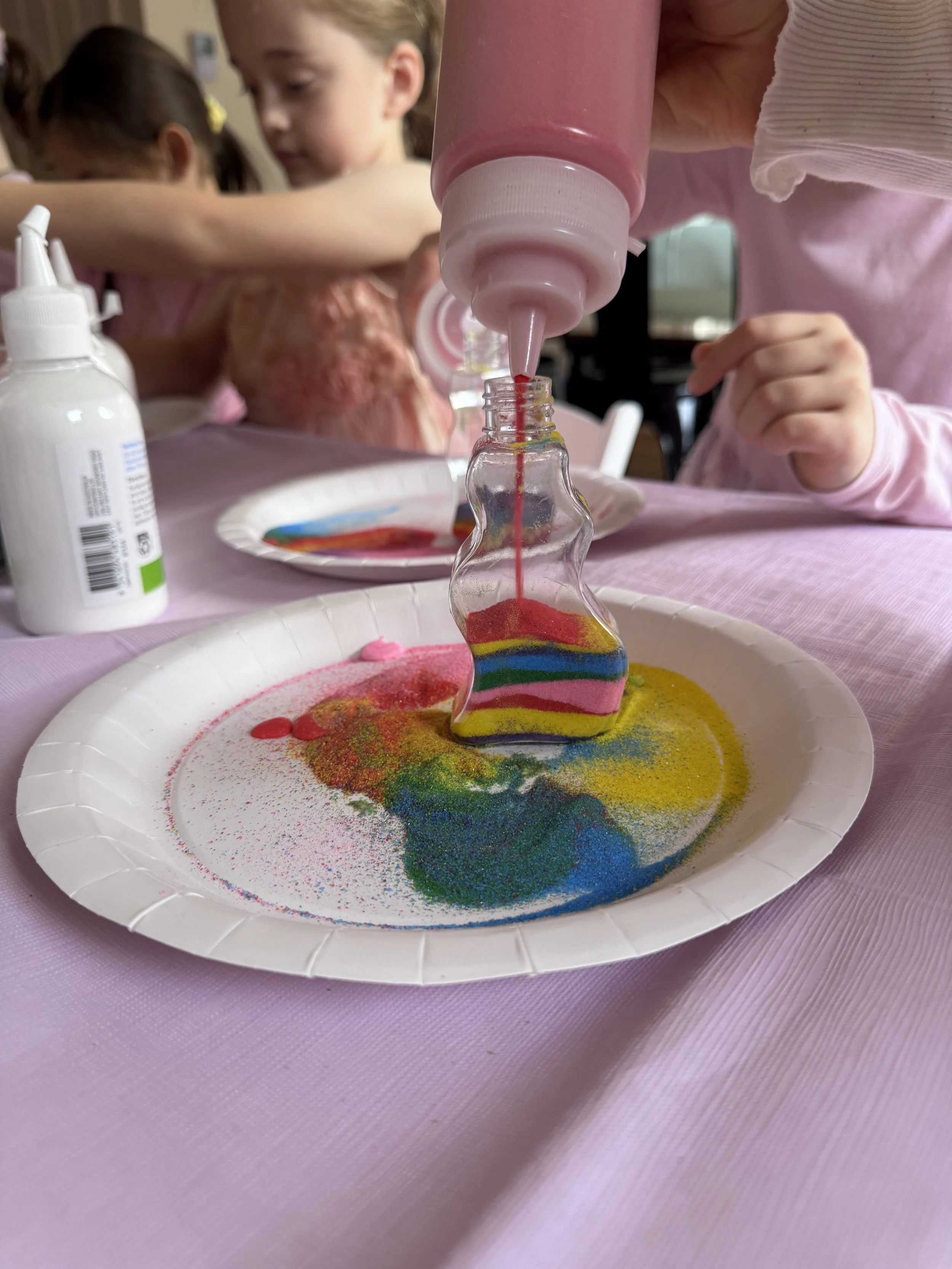 Children at a table creating rainbow-colored sand art, with a focus on a bottle dispensing colored sand into a shaped container on a paper plate. kids soiree craft activity parties