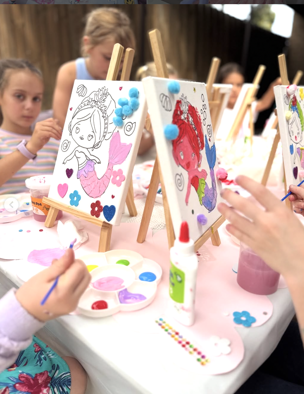 Children engaging in a painting activity at a table with small canvases on mini easels. The artwork features mermaids, some partially colored, surrounded by decorative elements like hearts and flowers. kids soiree craft activity parties