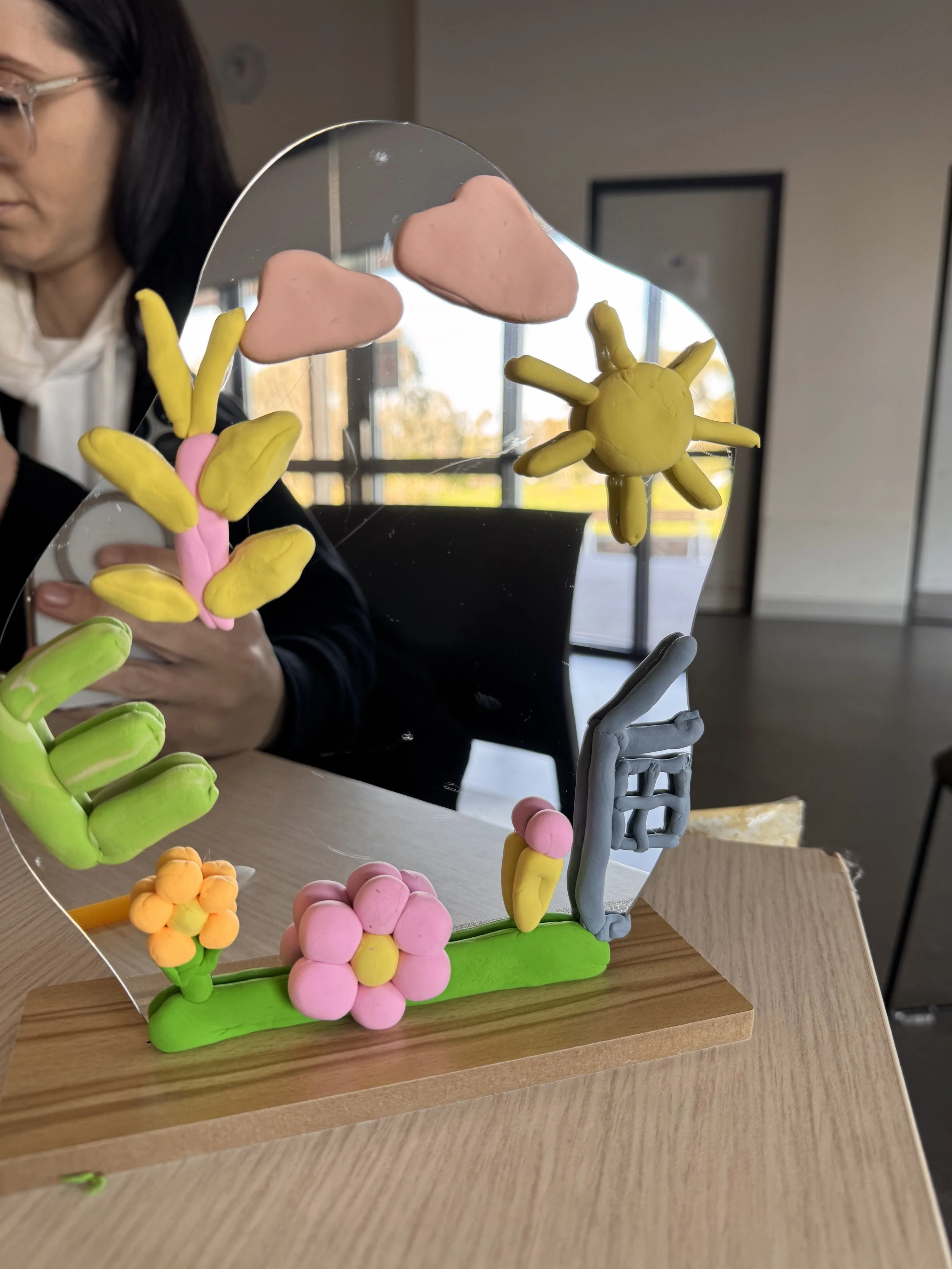 Colorful clay art of a sun, pink clouds, a flower, a hand, and a ladder on a mirror, with a woman in the background. kids soiree air clay party