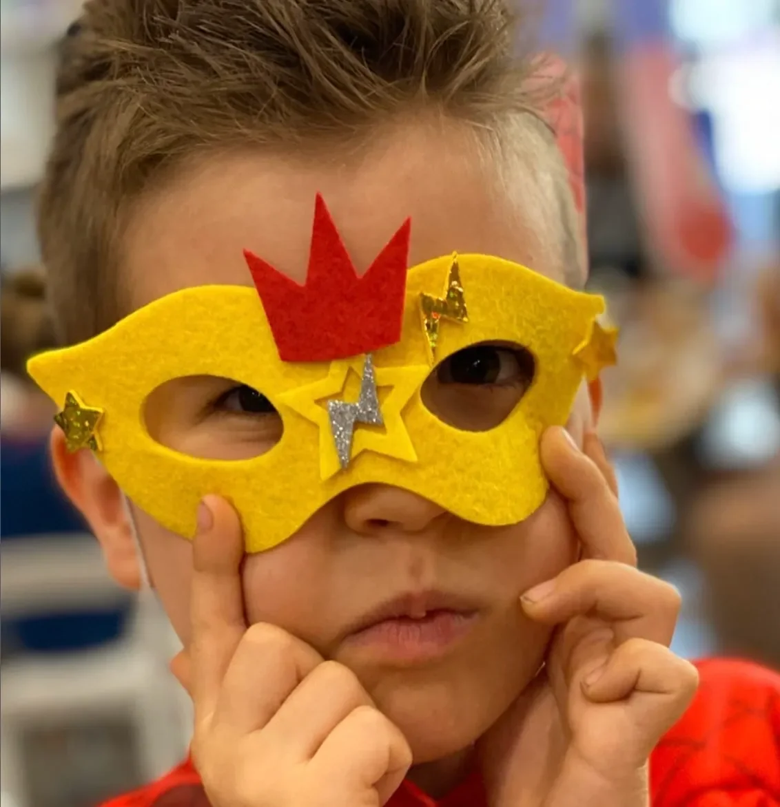 A young boy with light brown hair wearing a yellow superhero mask decorated with stars, a lightning bolt, and a crown. He is holding the mask with both hands, covering part of his face, and looking directly at the camera. kids soiree craft activity