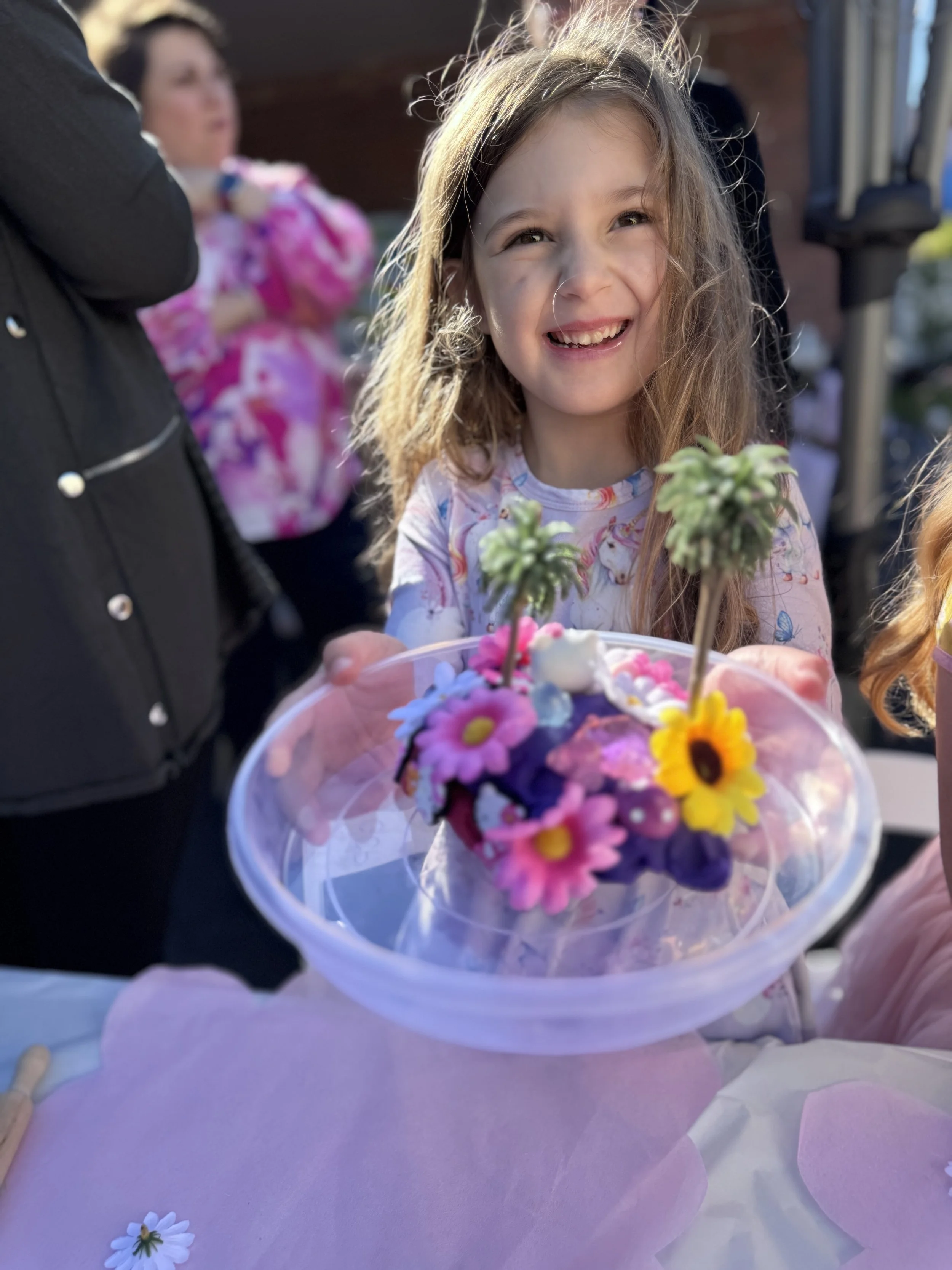 A young girl with long hair smiling while holding a bouquet of colorful flowers, with a blurred woman in a pink outfit in the background. kids soiree craft activity parties