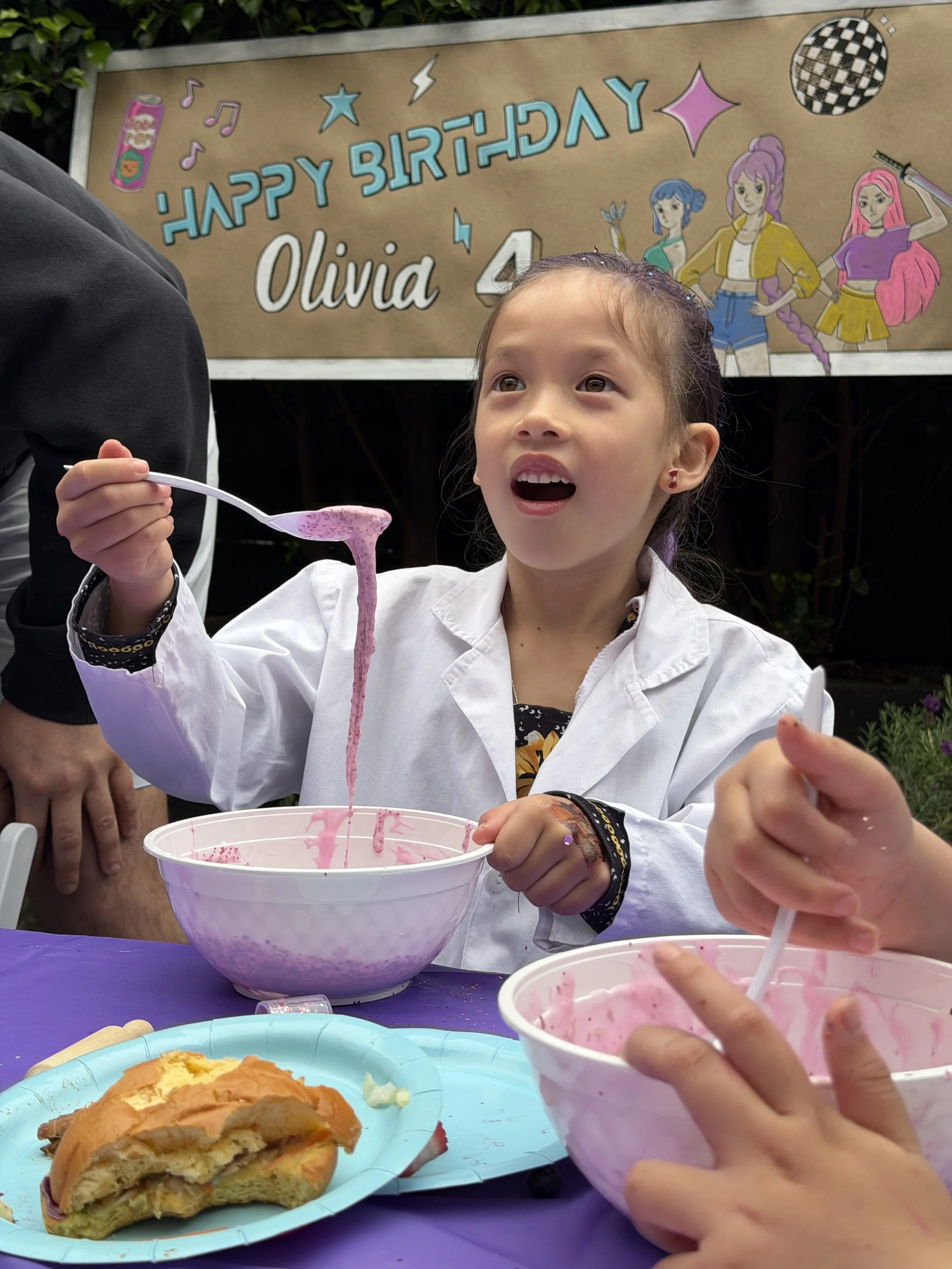 A young girl with a surprised expression sitting at a table with pink frosting on her spoon, a bowl of pink frosting in front of her, and a plate with a partially eaten piece of cake. In the background, a sign reads 'kids soiree craft activity partie