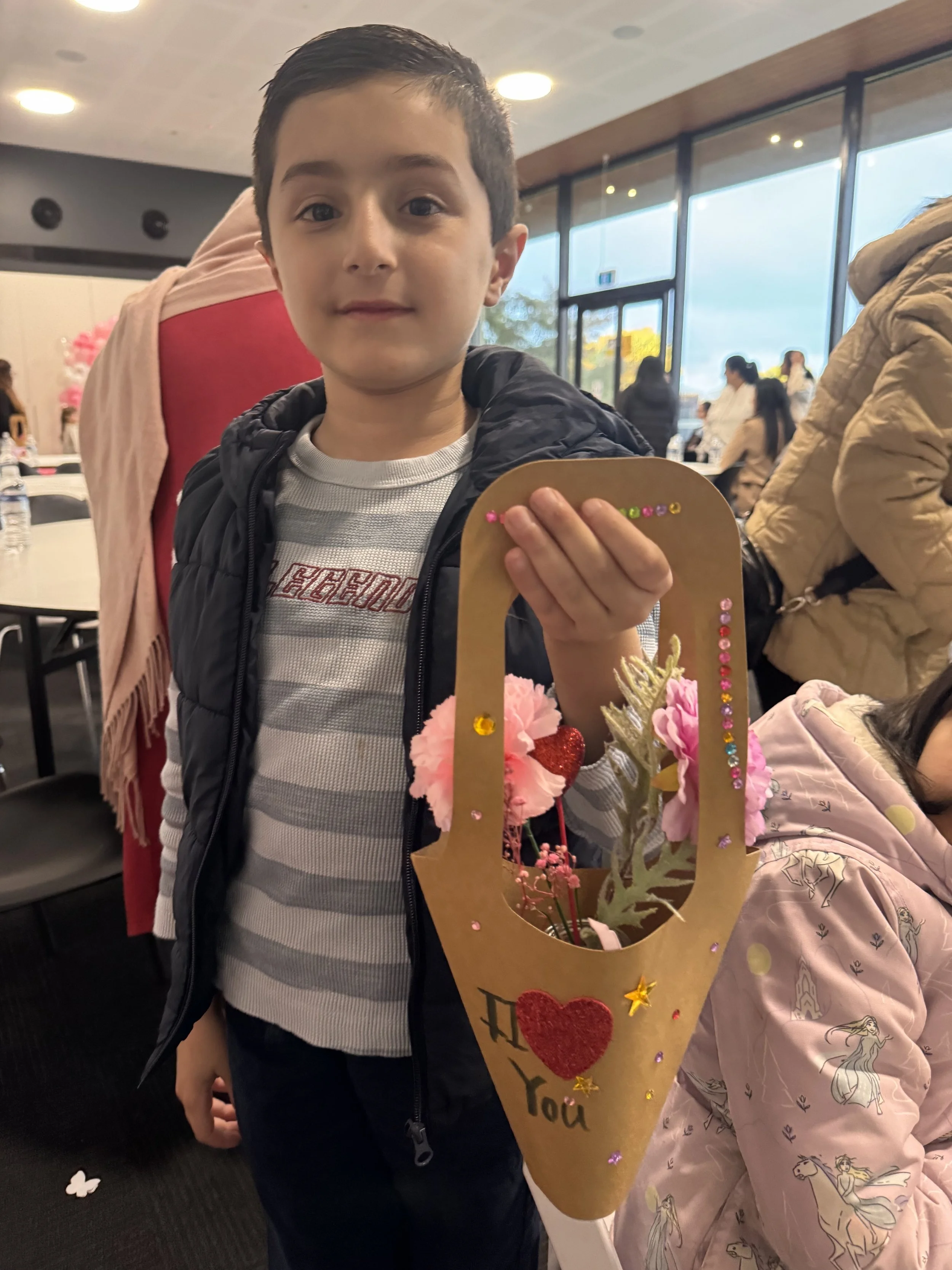 A young boy with short dark hair and wearing a striped gray and white shirt and a dark jacket holding a decorative card or banner with pink flowers, gold stars, and the words 'kids soiree craft activity parties