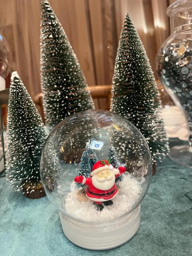 Christmas scene with a snow globe featuring Santa Claus, surrounded by three decorated miniature Christmas trees with snow-like accents, set on a green surface. kids soiree craft activity parties