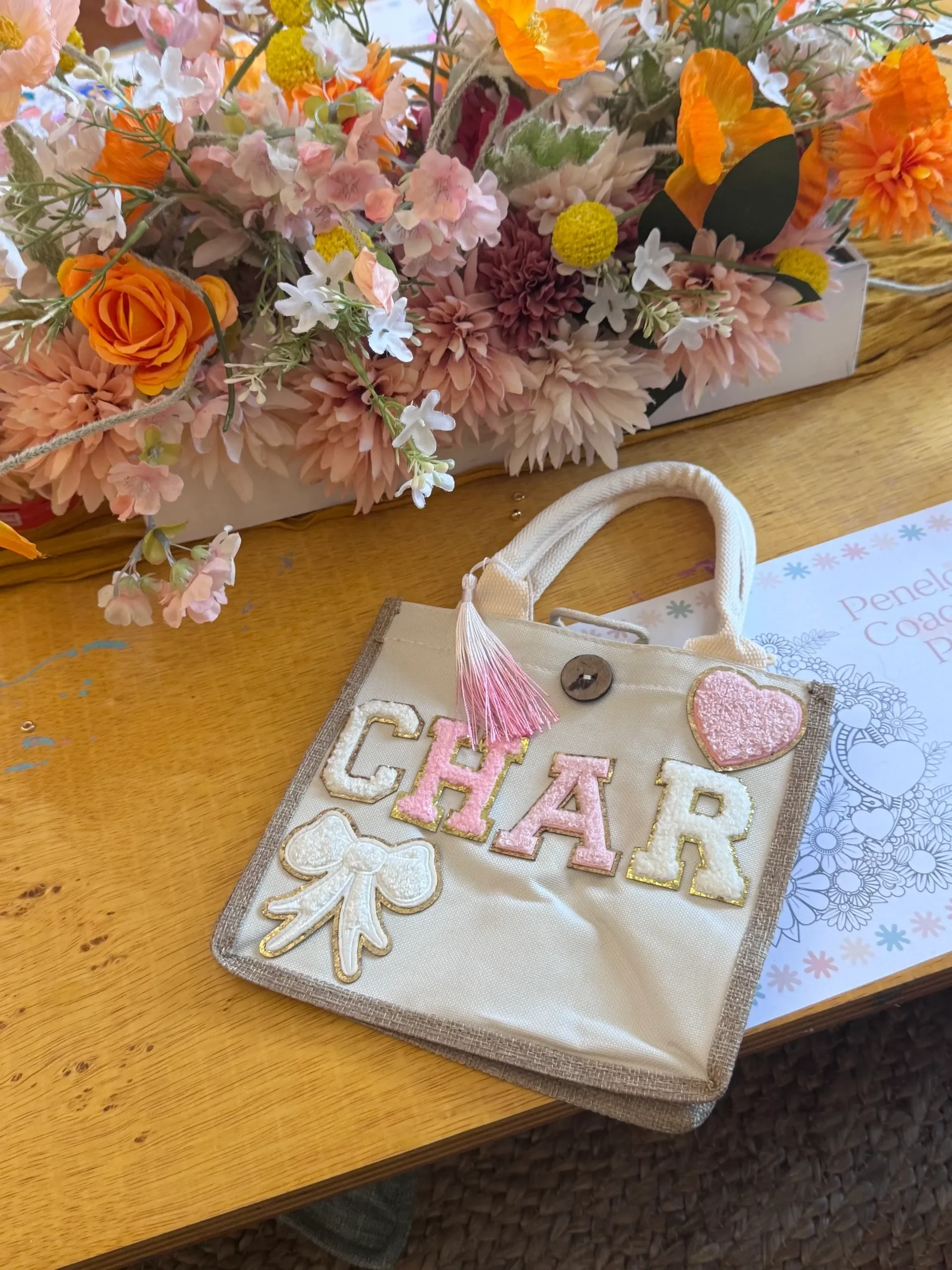 A beige fabric bag with embroidered pink, white, and gold letters spelling ''GIRL'' and a pink heart patch, along with white and gold embroidered flower and leaf designs, is placed on a wooden table. kids soiree craft activity parties