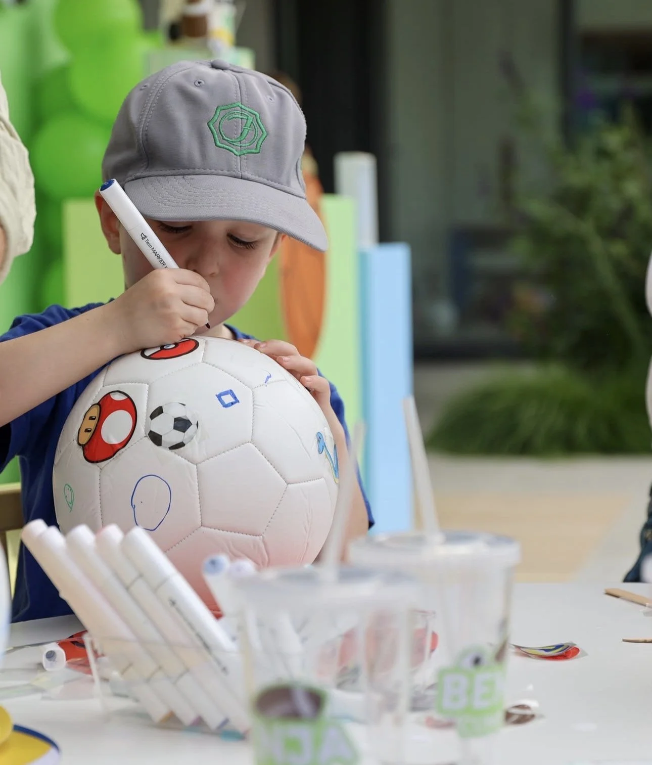 A young boy wearing a gray cap signs a decorated soccer ball with a black marker. The soccer ball has soccer-related images and doodles on it. The boy is sitting at a table with containers and markers, and there are kids soiree craft activity parties